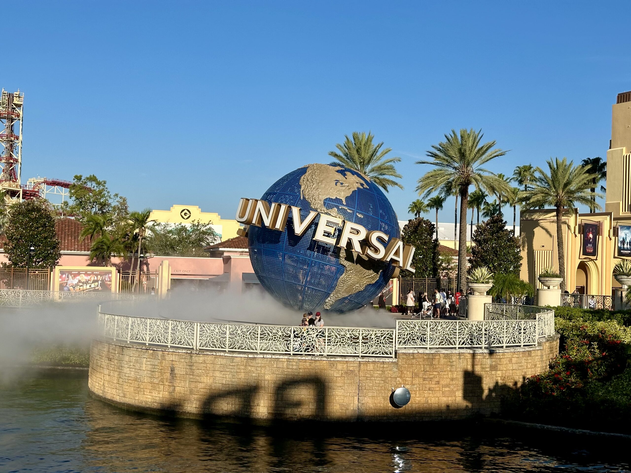 Large globe with "Universal" text, surrounded by mist, palm trees, and buildings in a theme park setting under a clear blue sky.