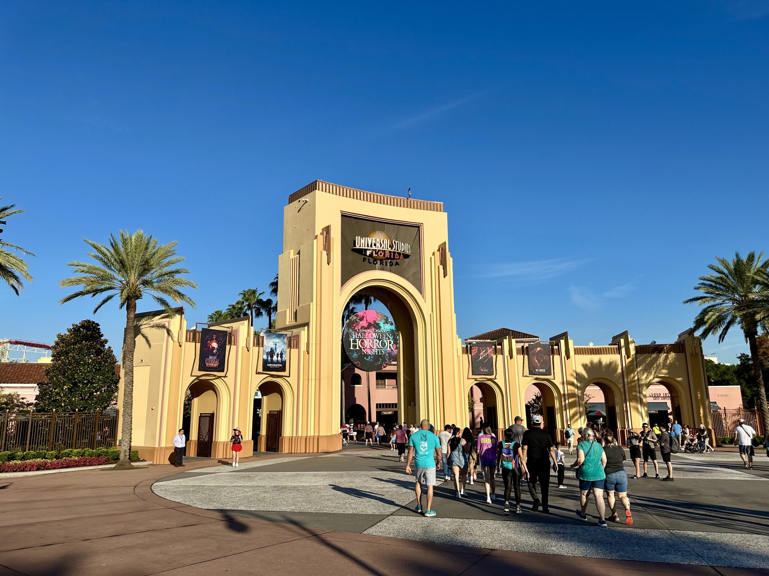 People walking toward the entrance of a theme park with palm trees and a clear blue sky.