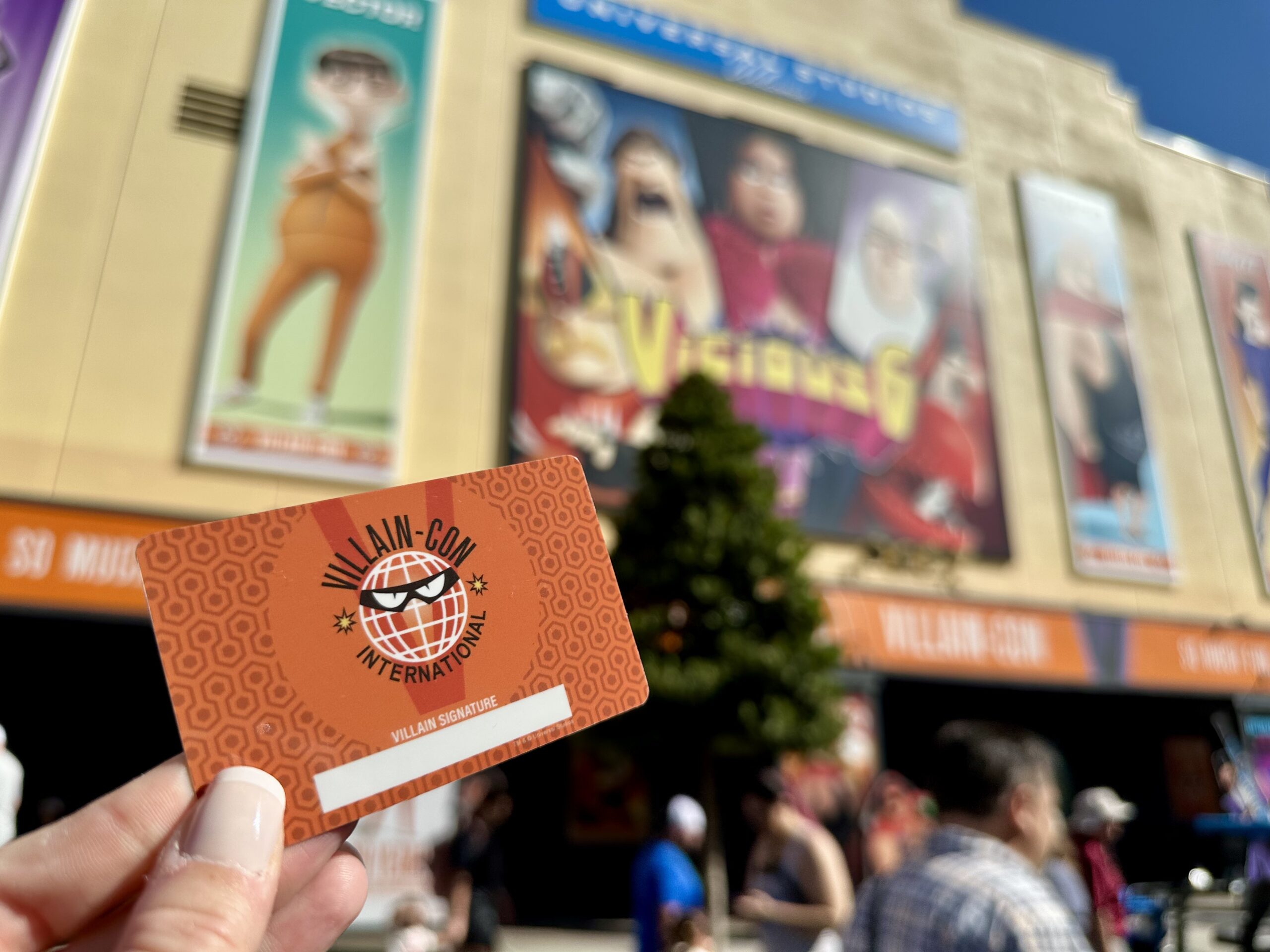 A hand holds a Villain-Con International card in front of a building with animated character posters and a crowd of people.