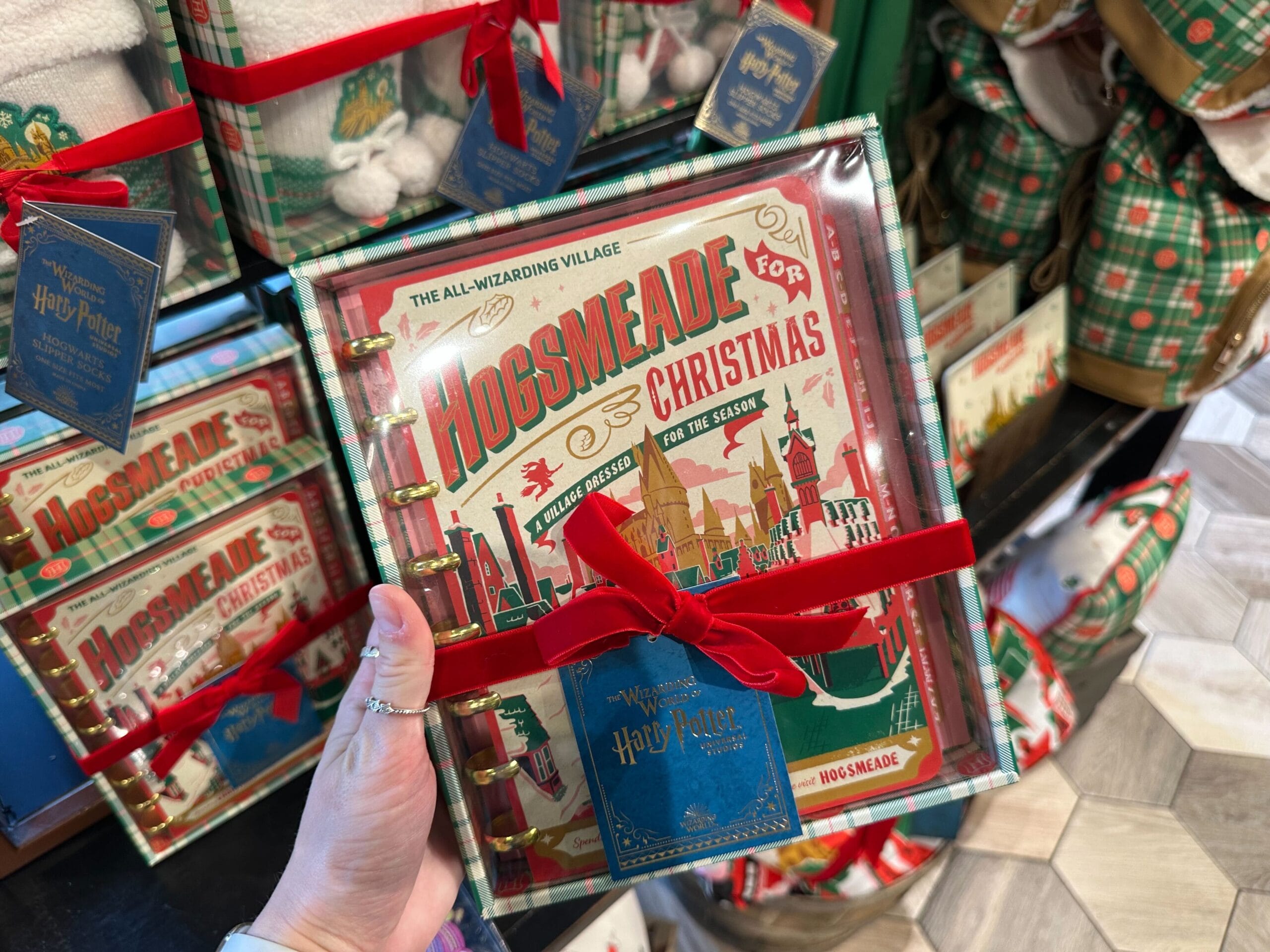 A person grips a Hogsmeade for Christmas book wrapped in a festive red ribbon, standing before shelves brimming with similarly enchanting treasures. A person grips a Hogsmeade for Christmas book wrapped in a festive red ribbon, standing before shelves brimming with similarly enchanting treasures.