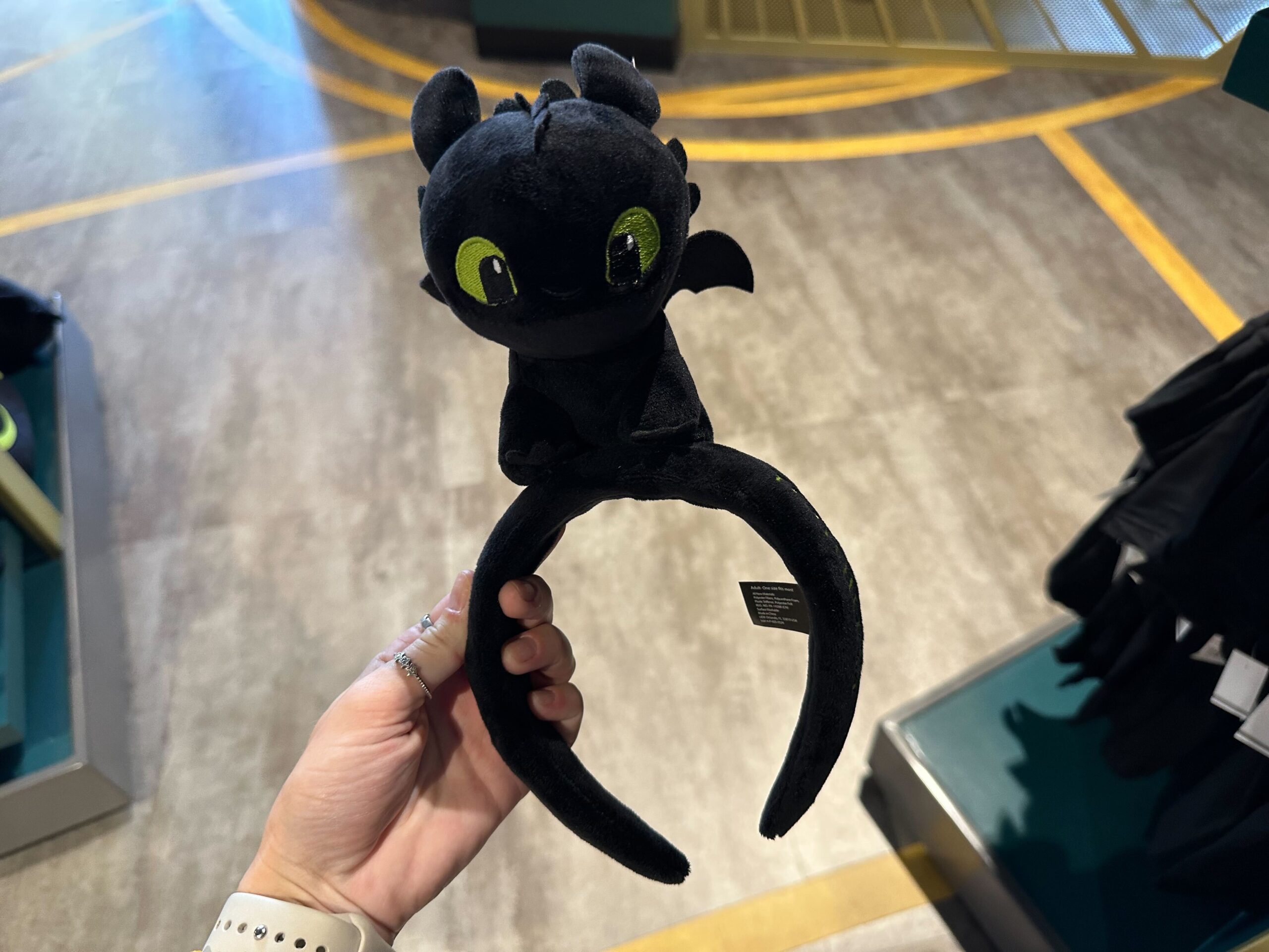 In a store setting, a person holds a new black plush headband featuring Toothless with green eyes, reminiscent of the magical charm found at Universal Orlando Resort.