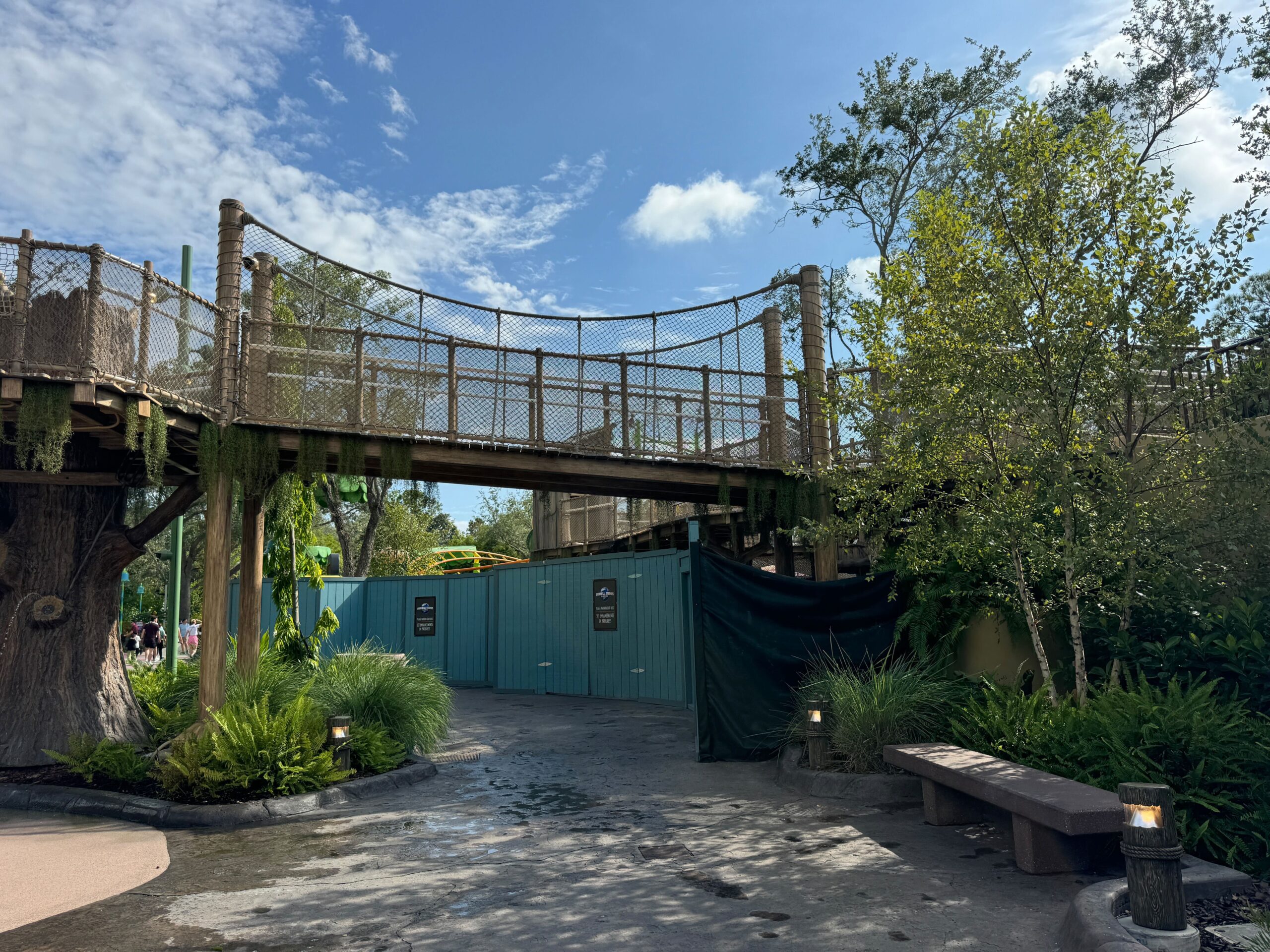 A wooden footbridge above a path resembles Shrek's Swamp, surrounded by lush trees and plants. Green construction barriers block the path, with a bench and the Little Ogres Playground pathway visible in the foreground, reminiscent of an enchanting corner at Universal Studios Florida.