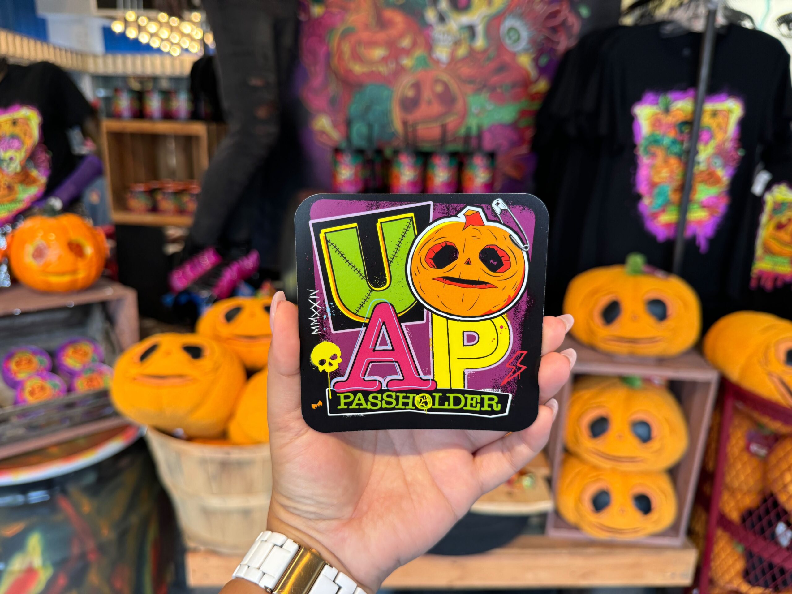 A hand holds a colorful UOAP passholder card in front of a display with various Halloween-themed items, including jack-o'-lanterns and t-shirts.