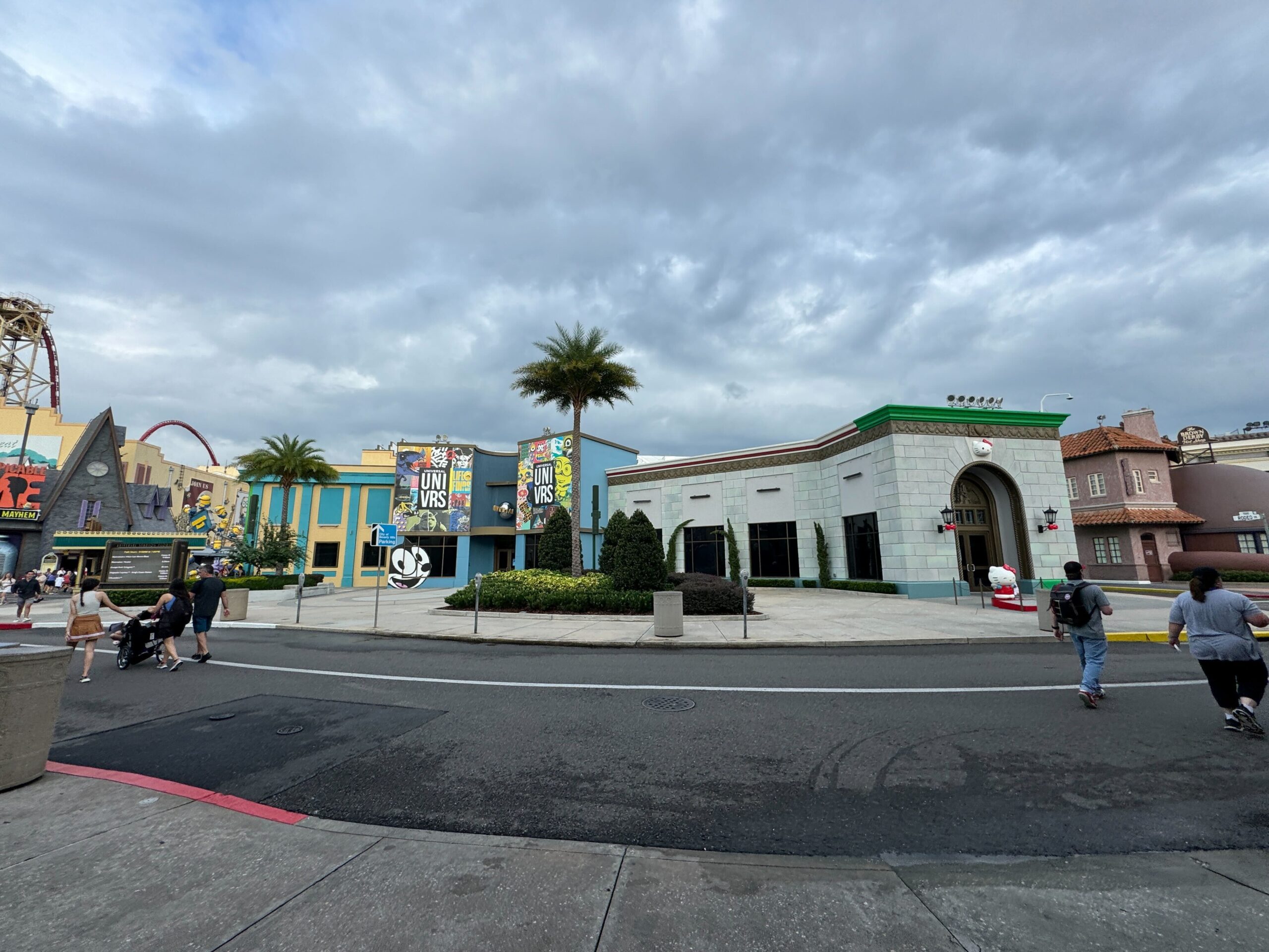 Amusement park street scene with several buildings, street lamps, palm trees, and cloudy sky. Visitors walk along the pathway.
