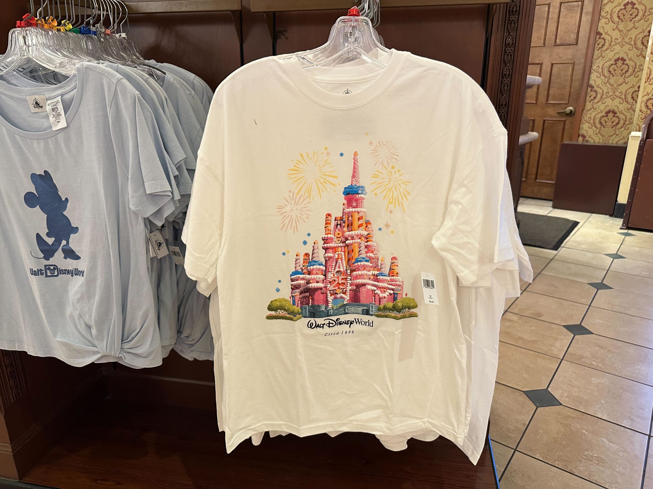 A white T-shirt adorned with a vibrant Anniversary Cake Castle design and "Walt Disney World" text, hanging alongside other shirts on a rack.