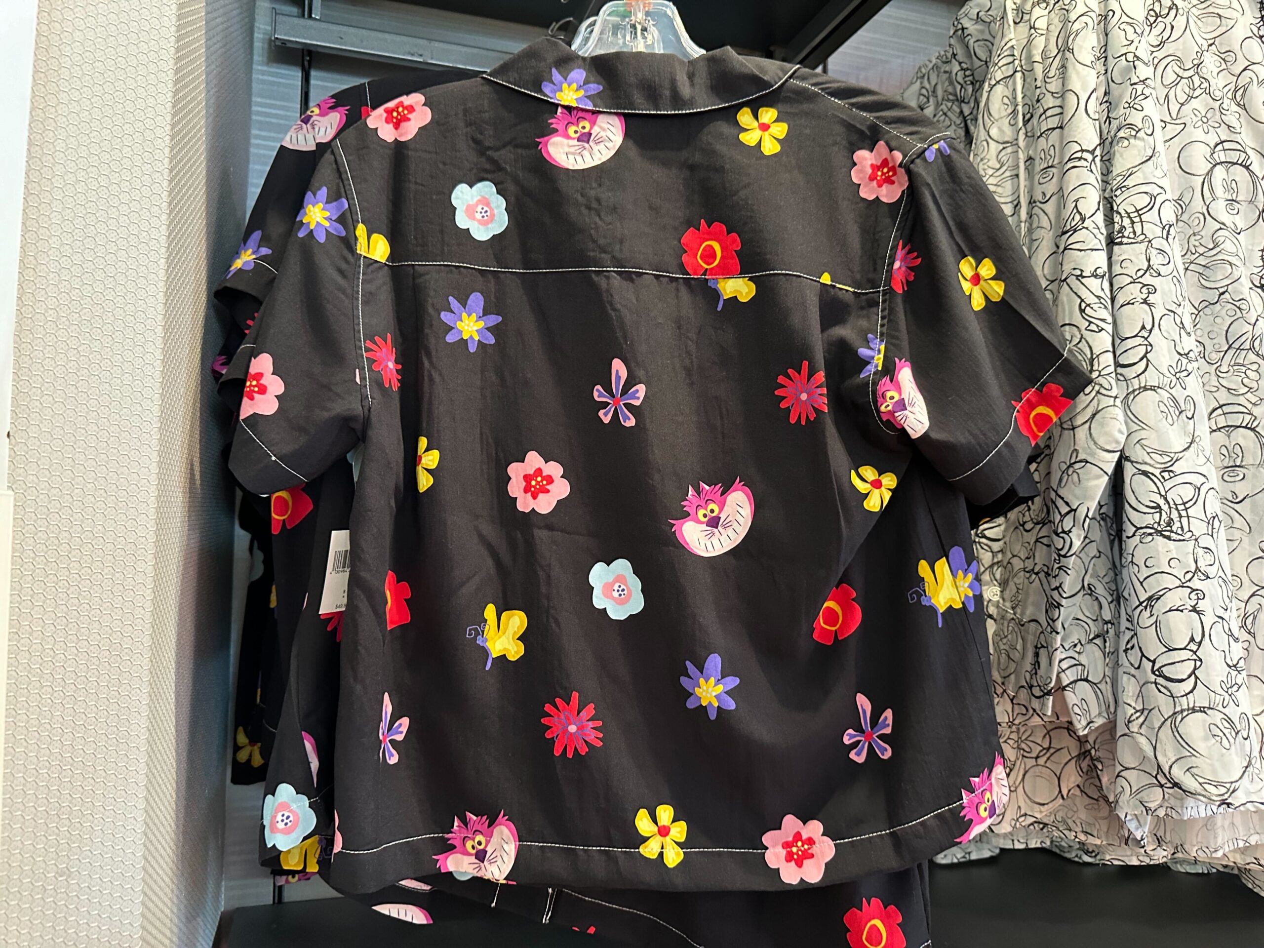 A colorful, floral-patterned black short-sleeve shirt reminiscent of an Alice in Wonderland adventure hangs on a rack.