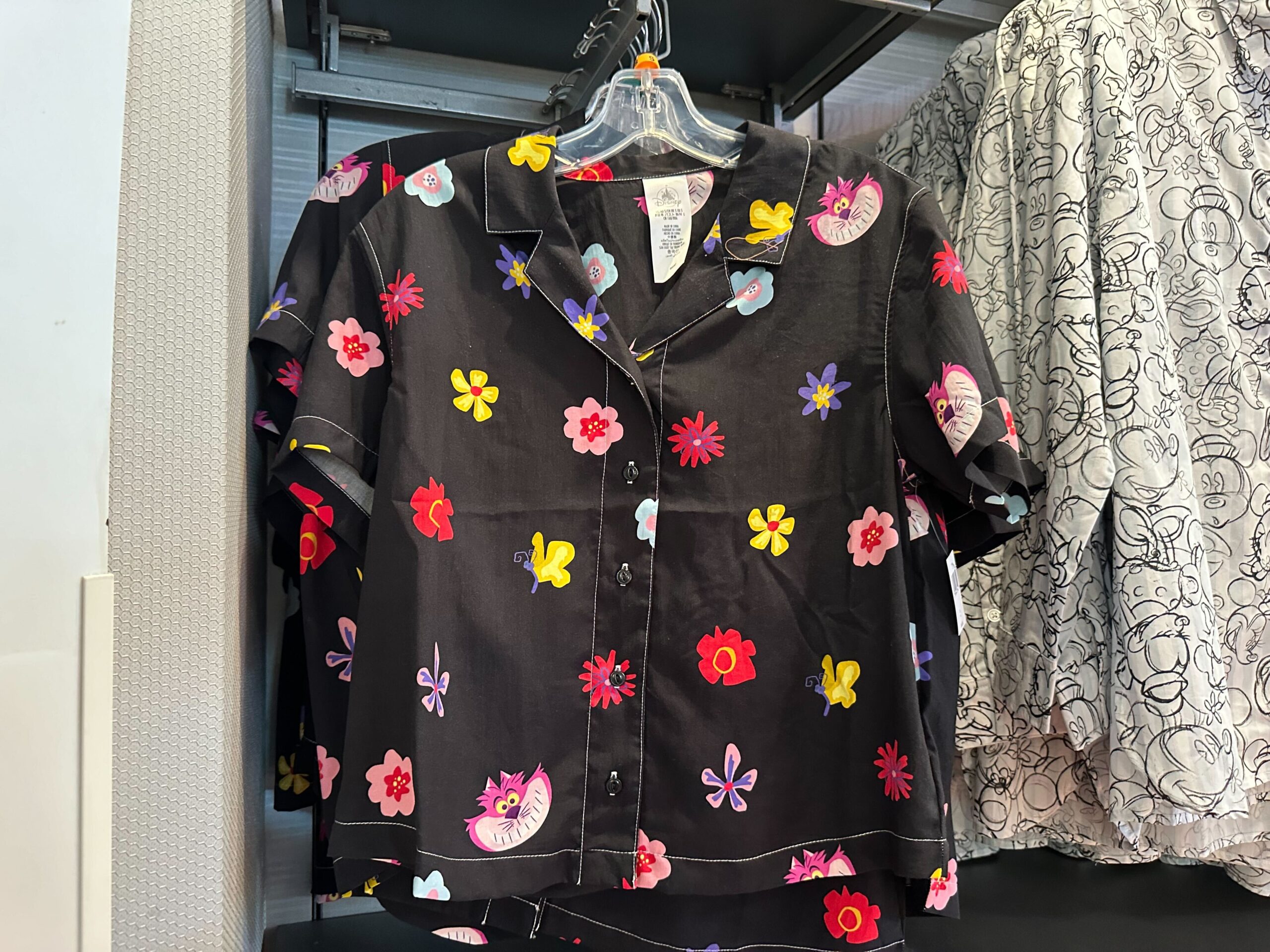 A black button-up shirt with colorful floral and cat designs reminiscent of Alice in Wonderland Merchandise hangs elegantly on a clothing rack.