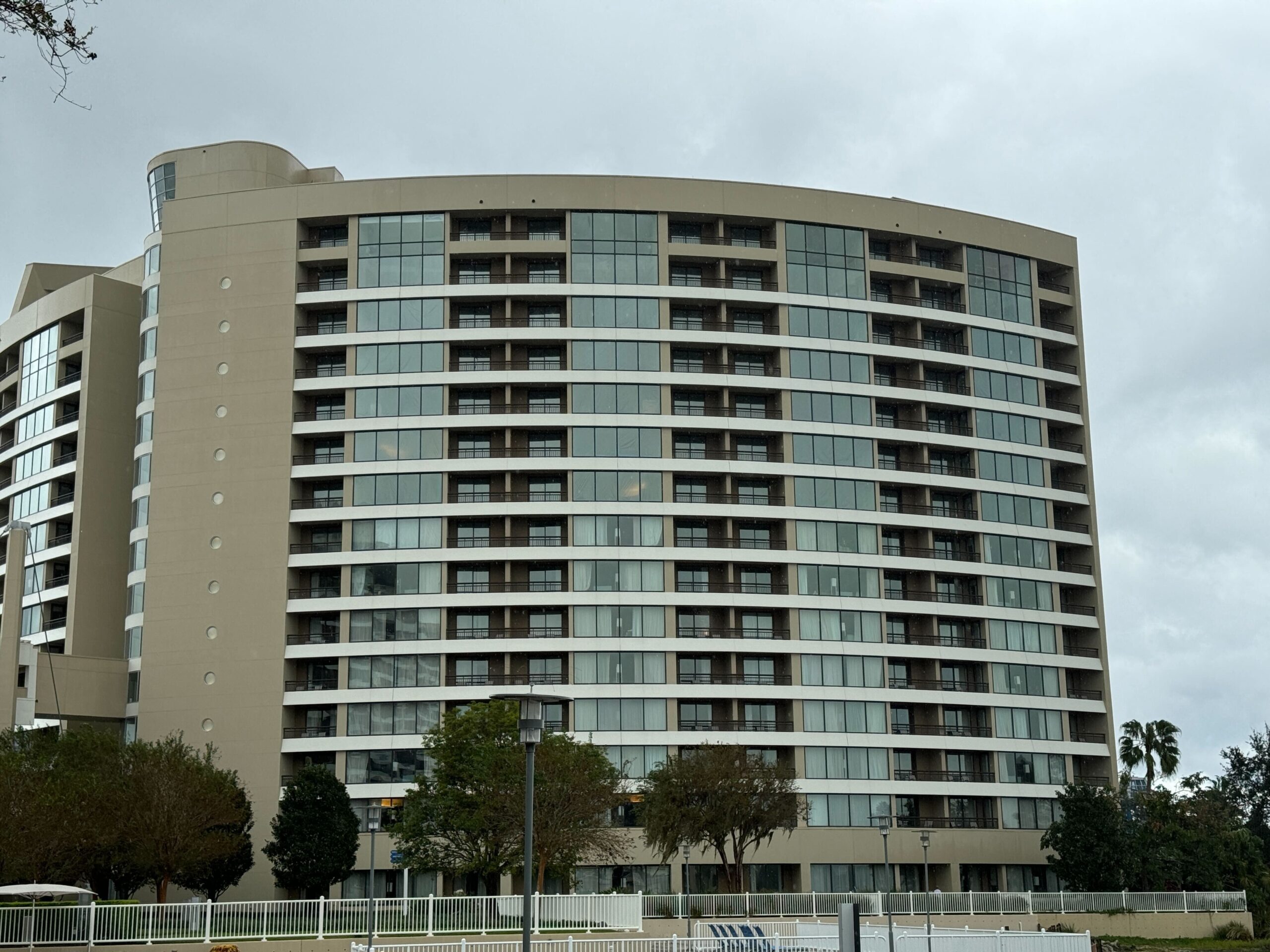A modern, multi-story hotel building with a curved design and glass windows. Trees and a fence are in front.