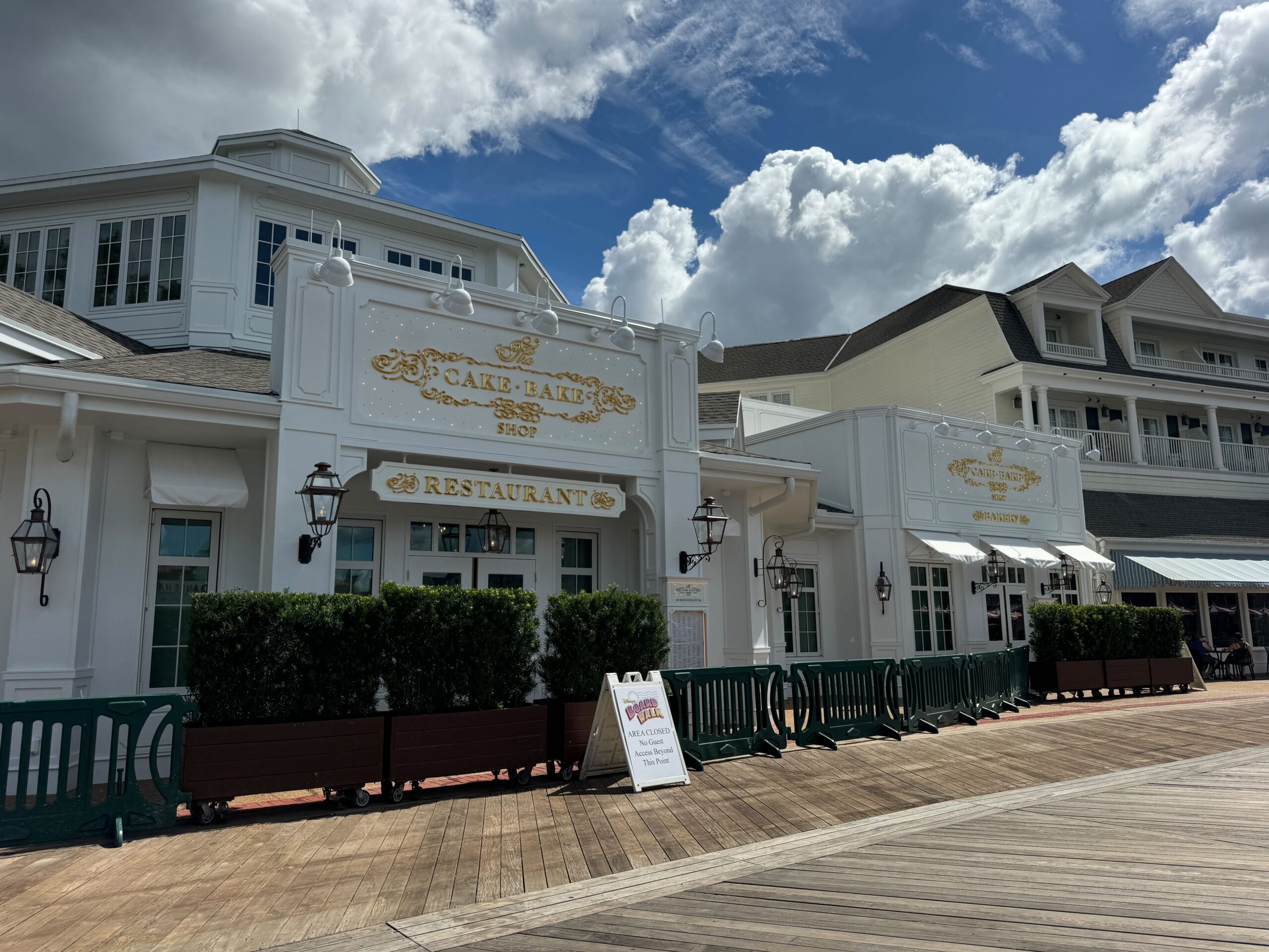 A white building adorned with signs for a Cake Bake Shop and restaurant stands proudly. Surrounded by lush greenery, it features a charming wooden boardwalk out front. Under the cloudy sky, the inviting menu promises delightful treats within.