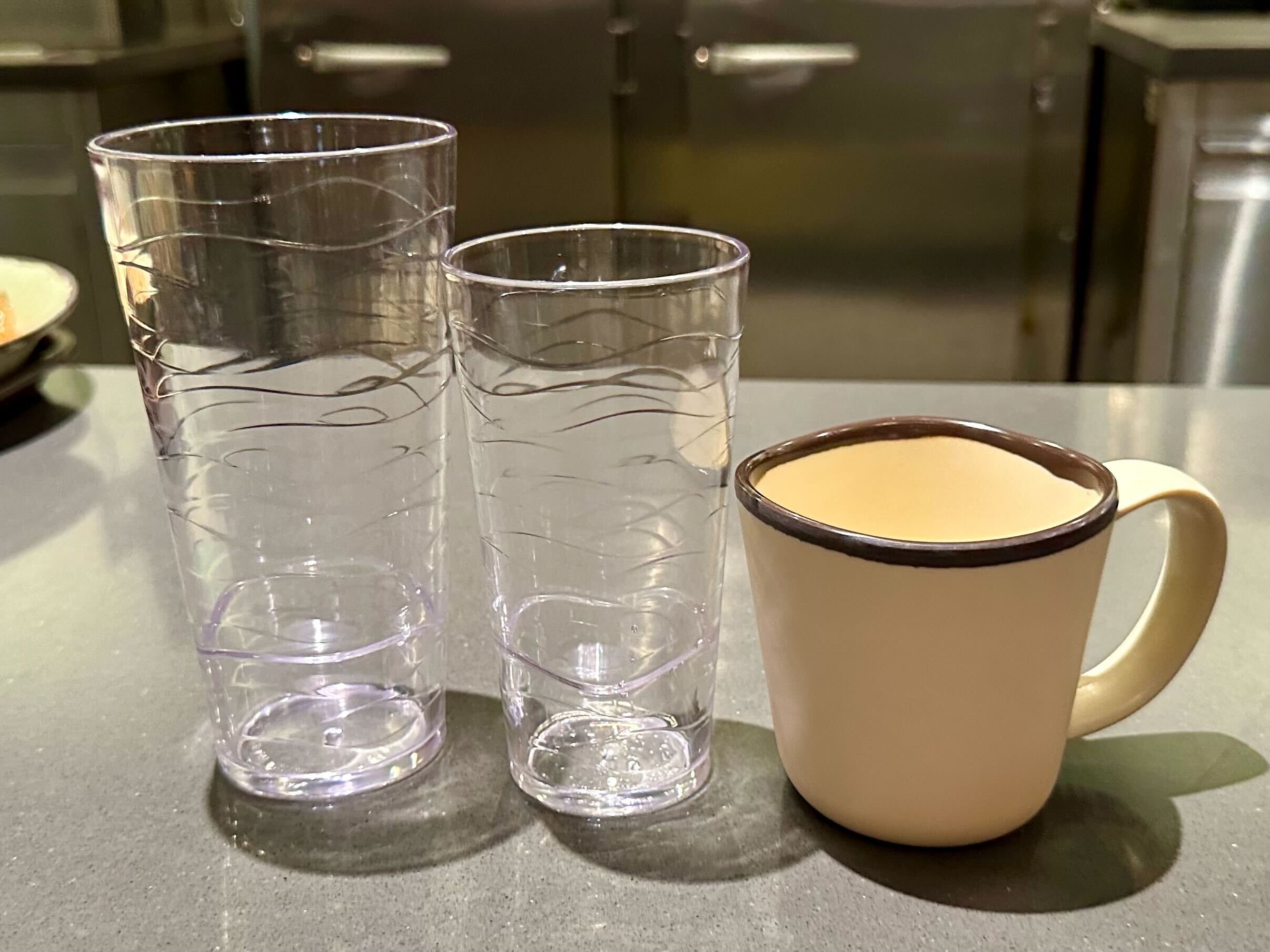 Three empty drinking vessels on a countertop: two clear, textured plastic cups and one beige ceramic mug with a brown rim.