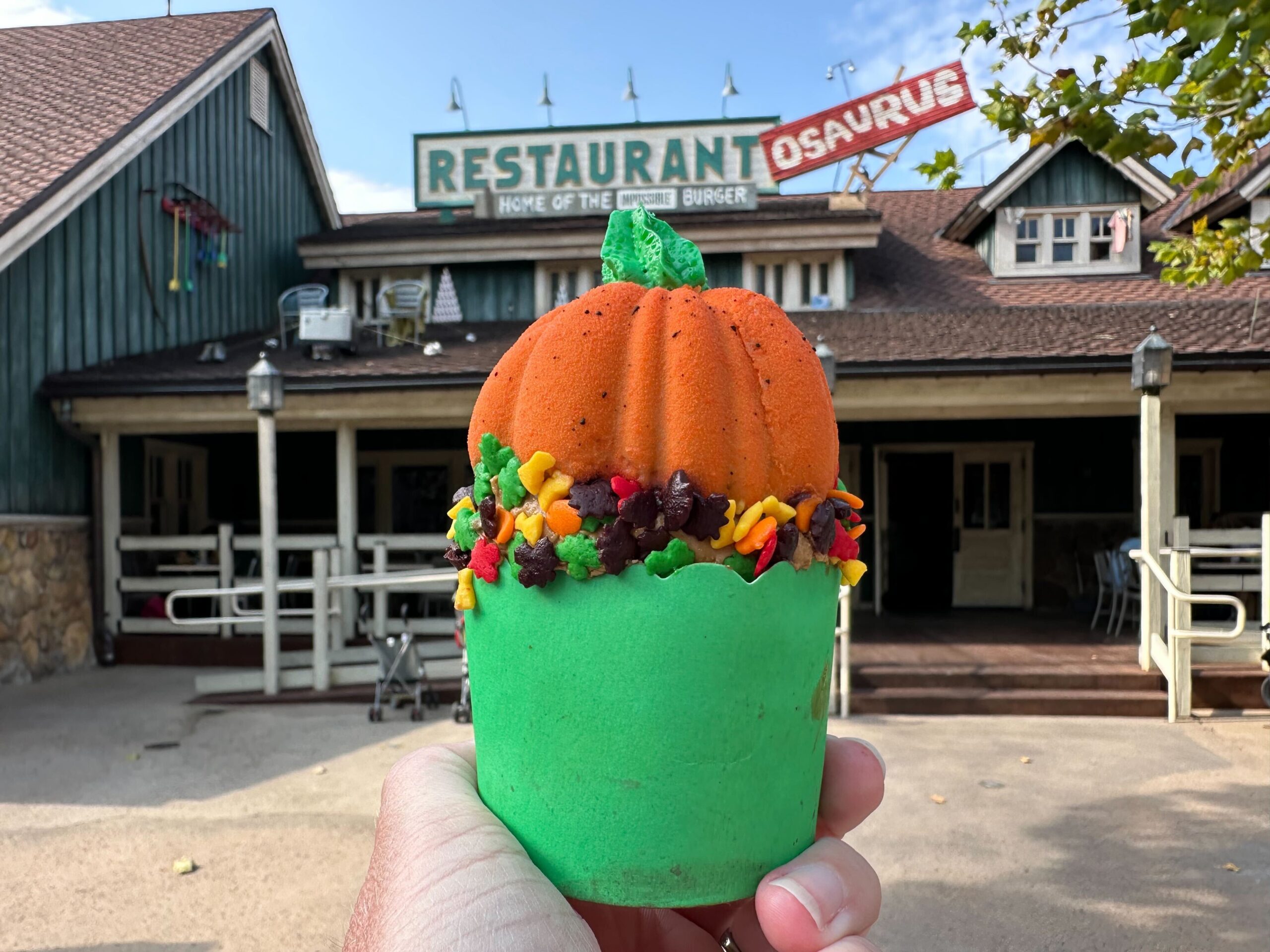 A hand holds a pumpkin-themed cupcake with green frosting in front of a rustic restaurant with a sign reading "Restaurant-osaurus.