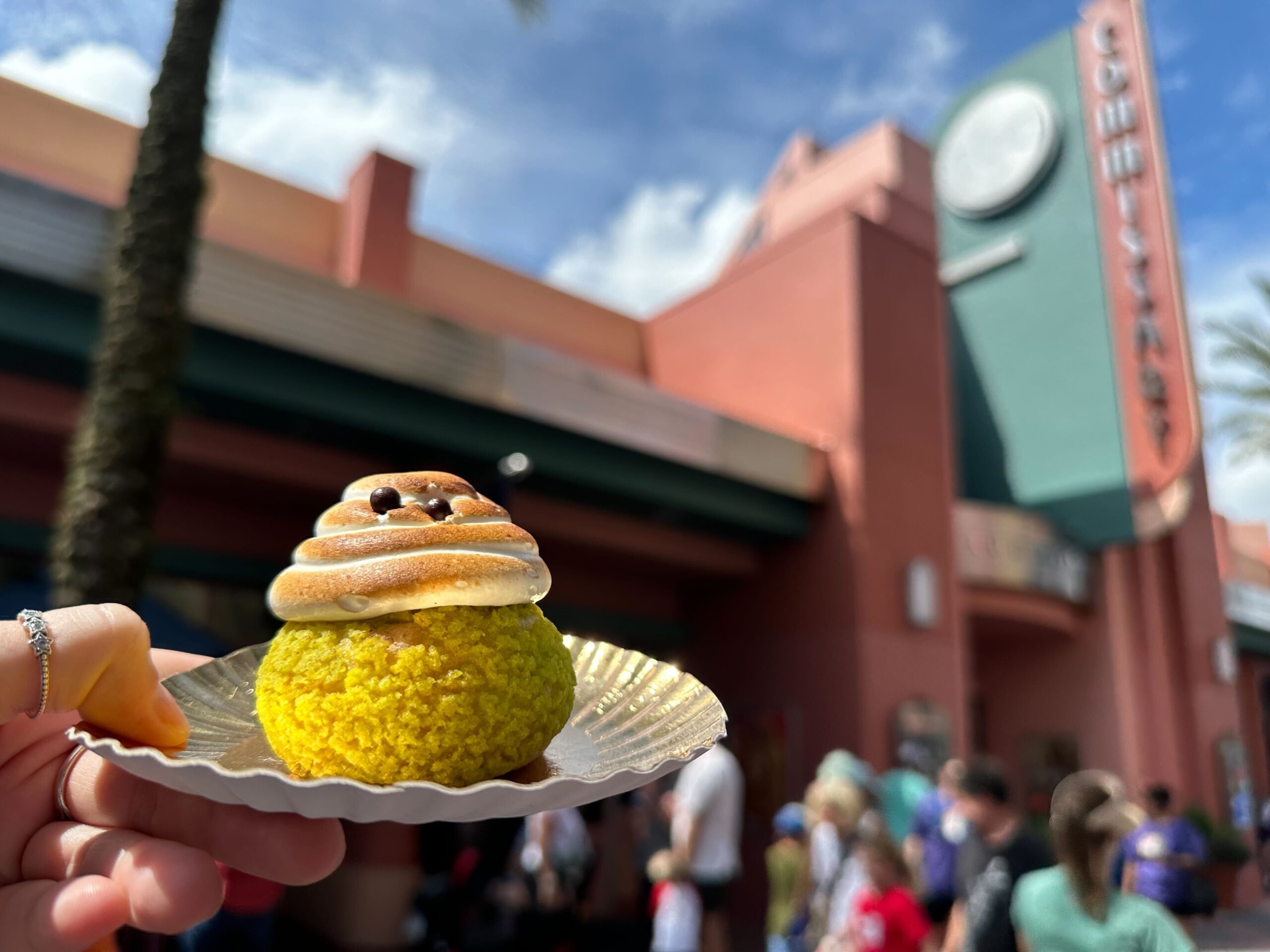 A hand holds a small dessert topped with meringue and chocolate chips in front of a retro-style building with a sign reading “Ghirardelli” and a crowd of people.