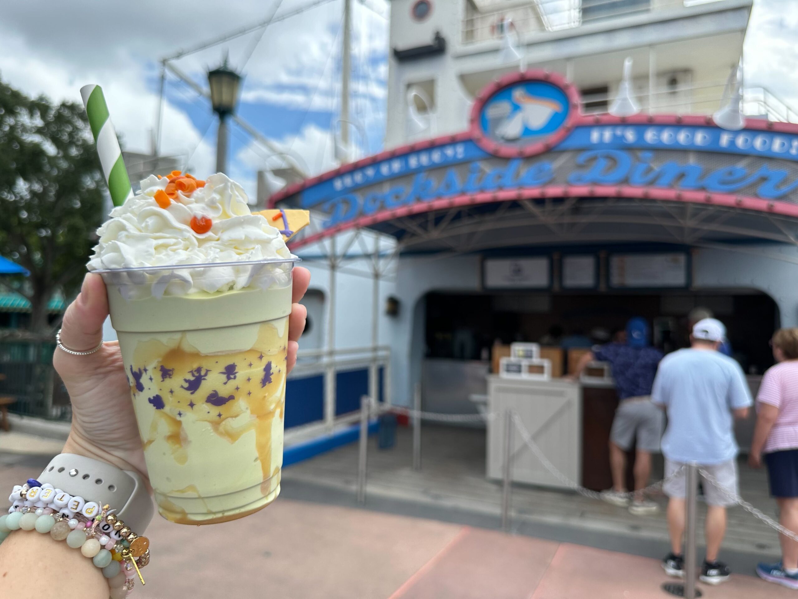 A hand holds a whipped-cream topped milkshake with a green straw in front of Dockside Diner. People are standing in line at the diner.