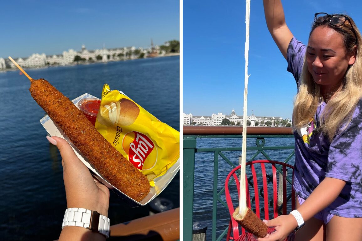 A person holds a corn dog with cheese pulling apart, next to a bag of chips and ketchup, with a waterfront view in the background.