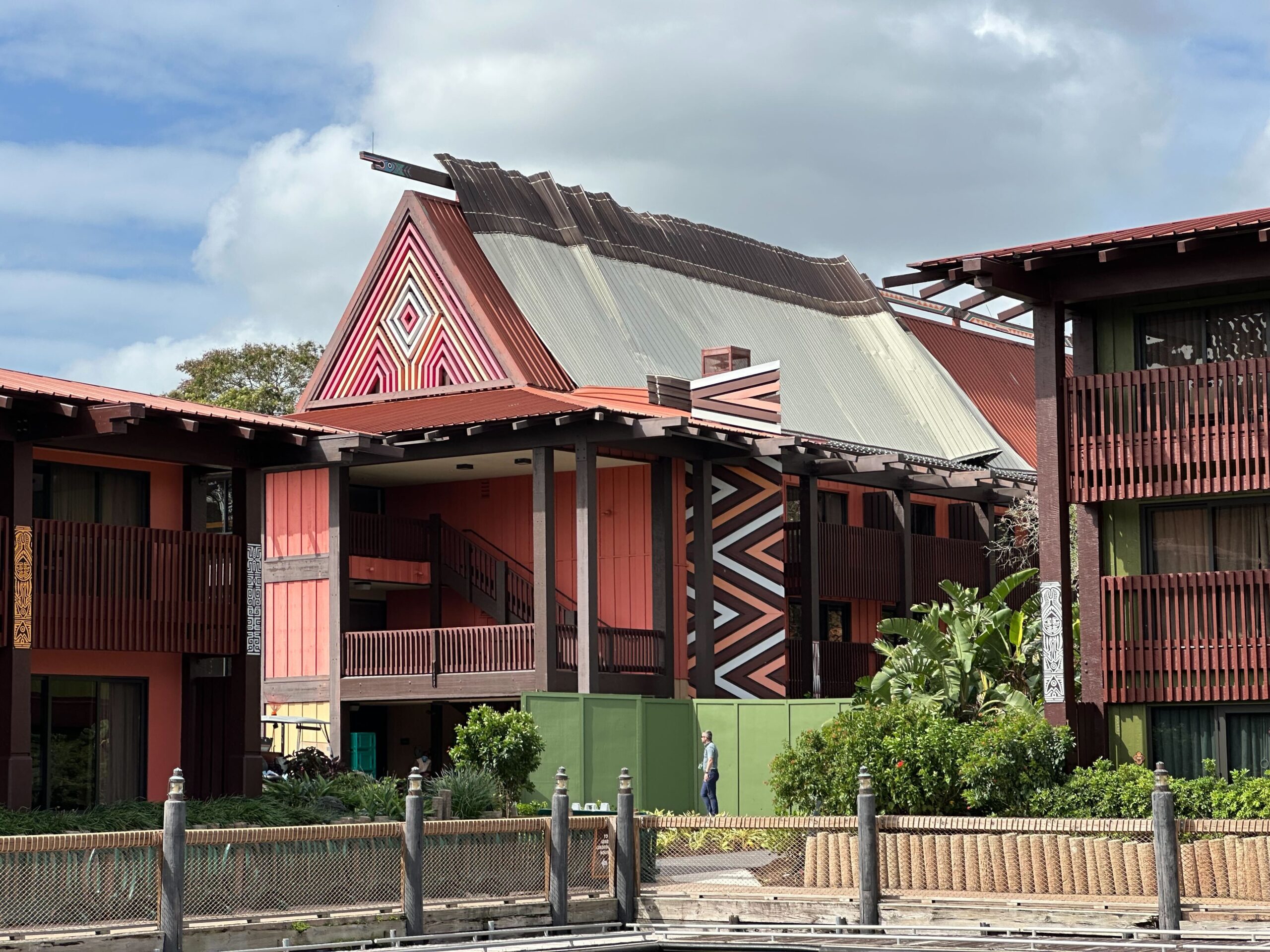 A building with geometric patterns and a pitched roof, surrounded by greenery, under a cloudy sky.