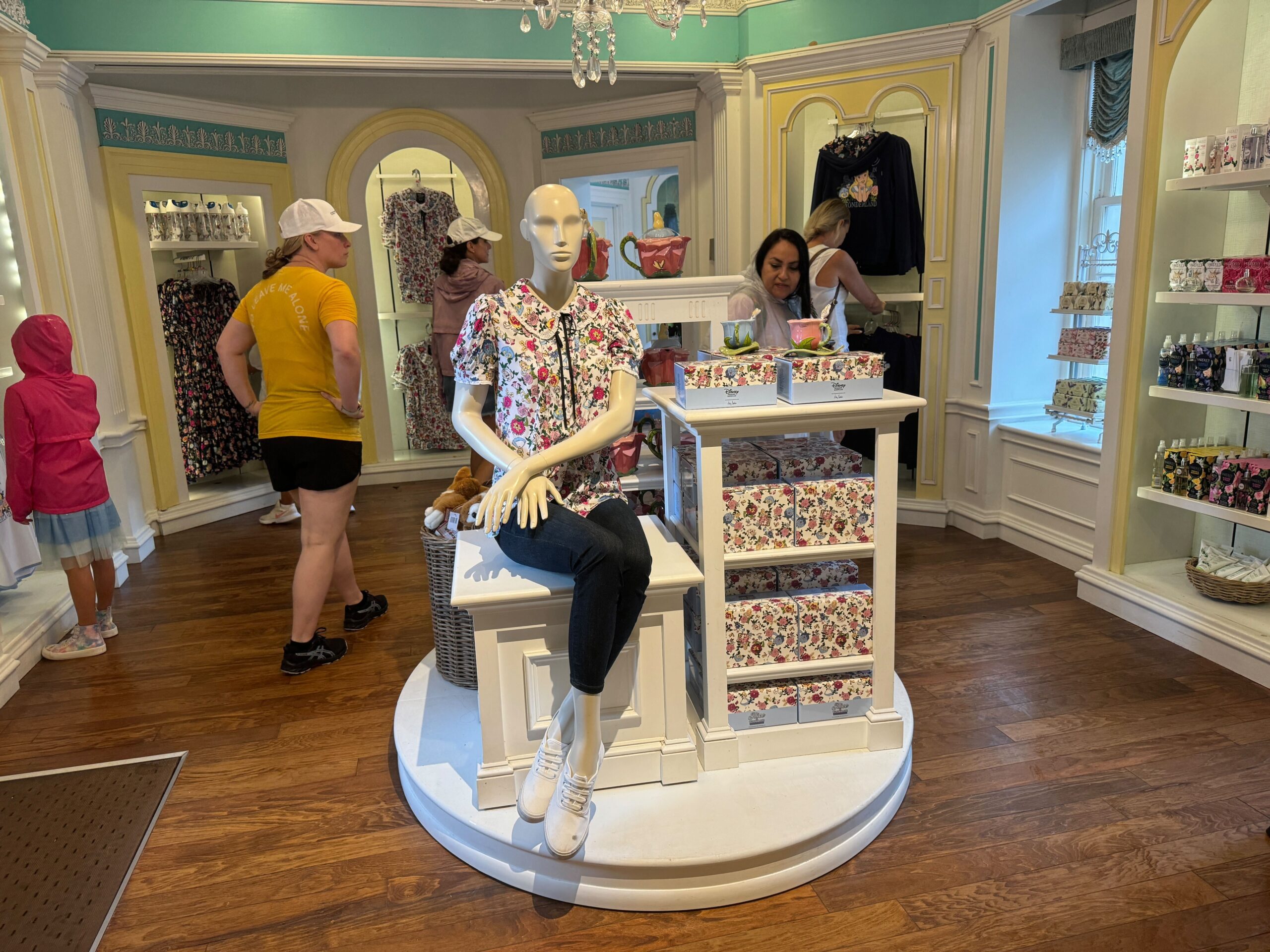 A mannequin in a floral shirt sits on a display stand in a clothing store. People shop around shelves filled with similar floral items.