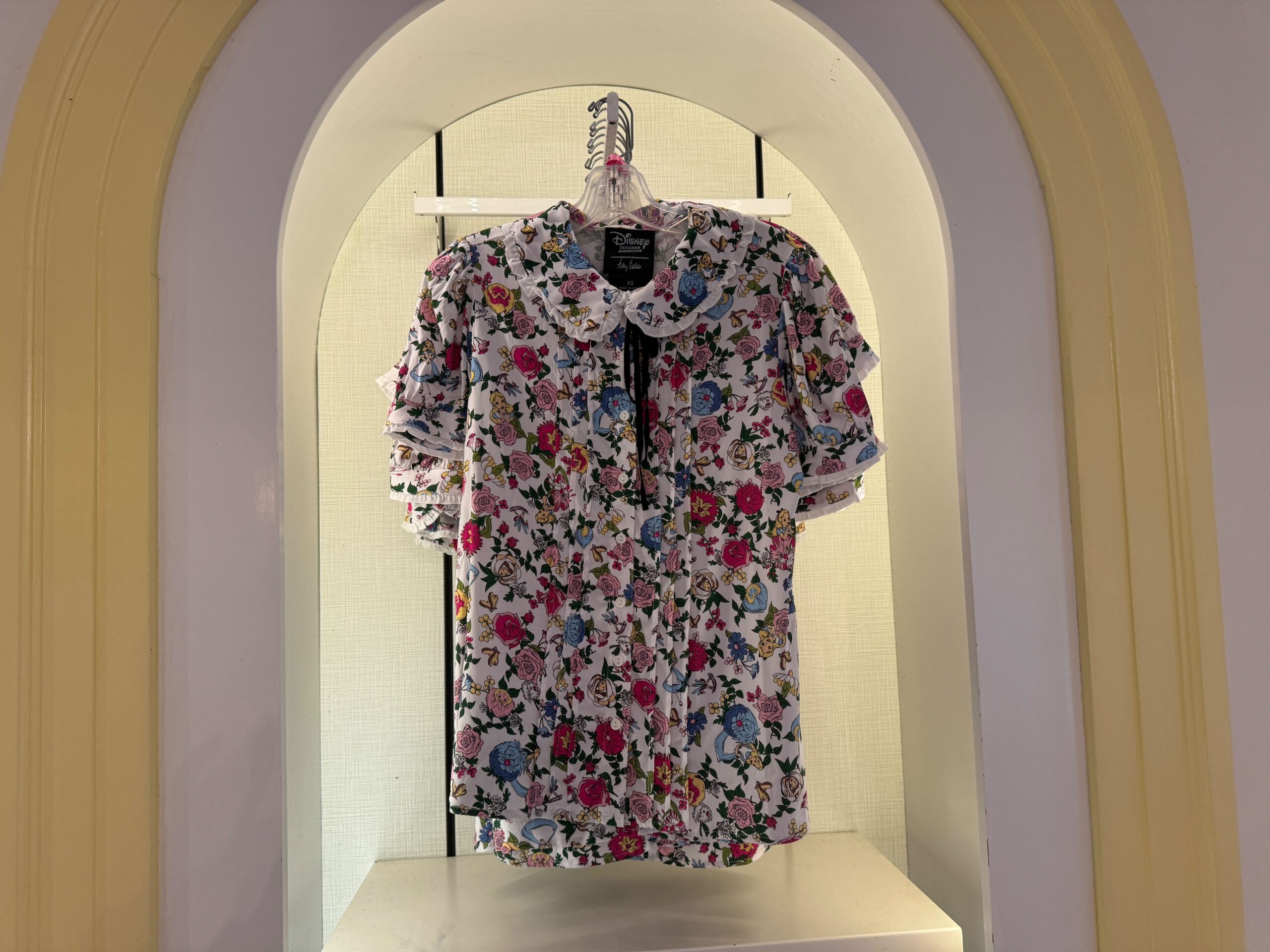 Floral print blouse with a ruffled collar displayed on a hanger in a lit alcove.