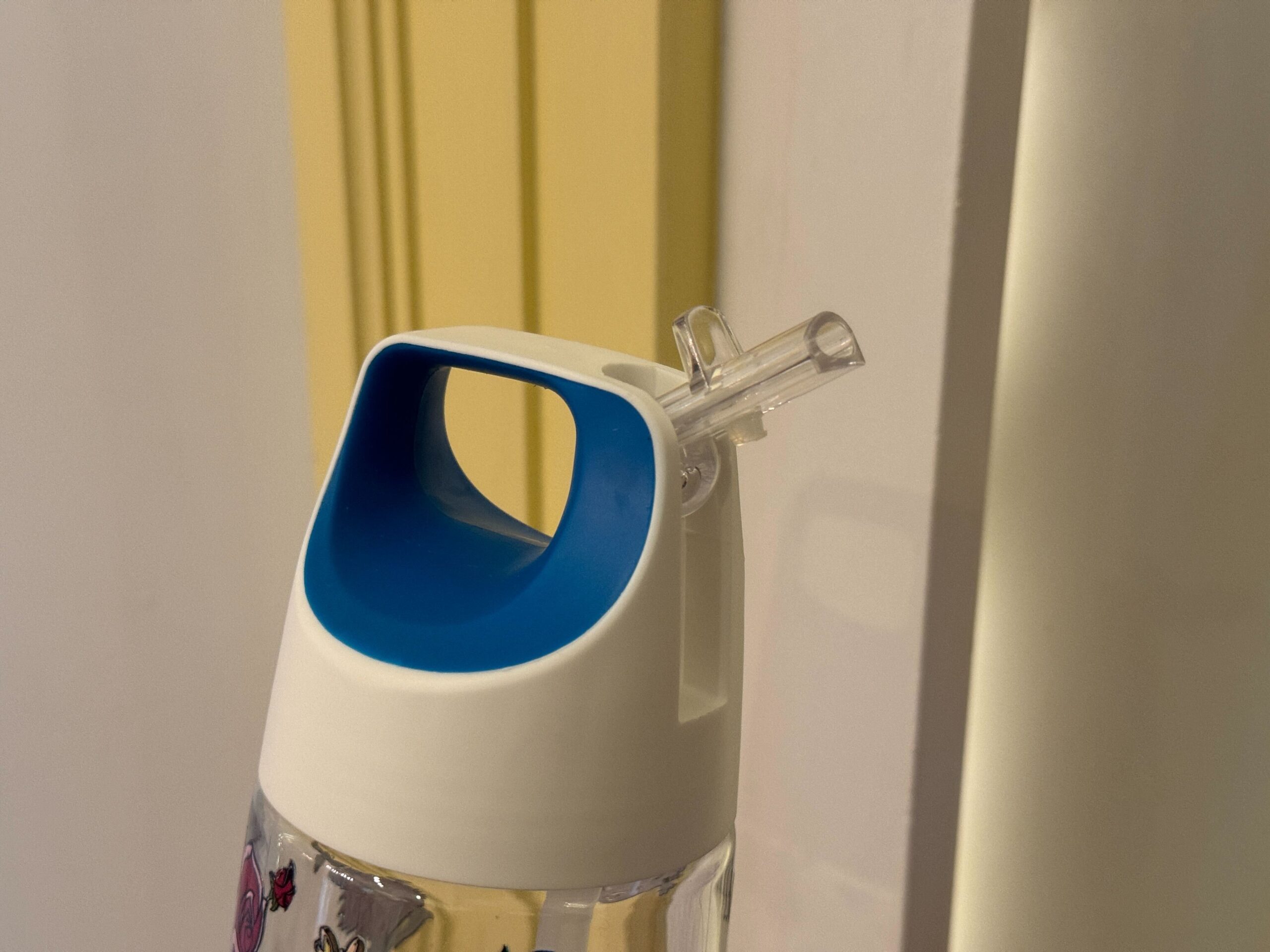 A close-up of a plastic water bottle with a blue and white lid, featuring a pop-up straw against a yellow and white painted background.