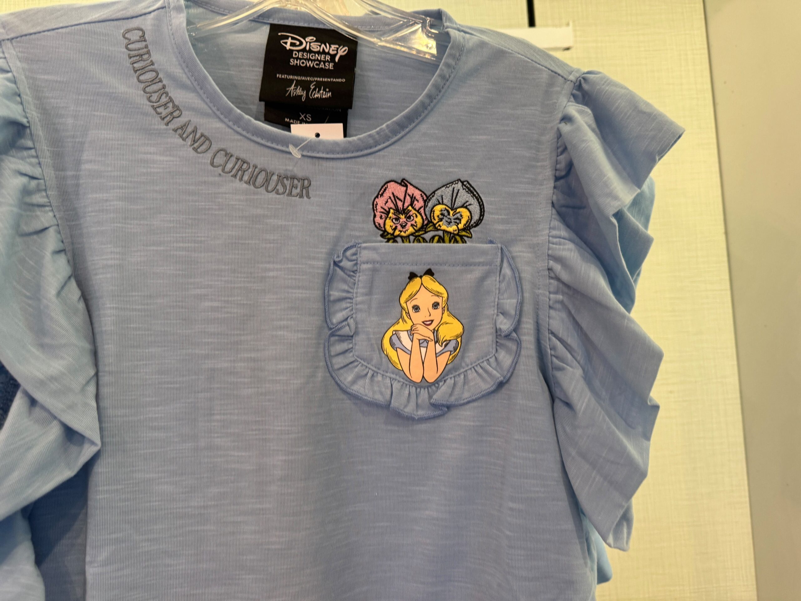 Light blue shirt with "Curiouser and curiouser" text. Features illustrations of flowers and a girl with blonde hair on the ruffled pocket.