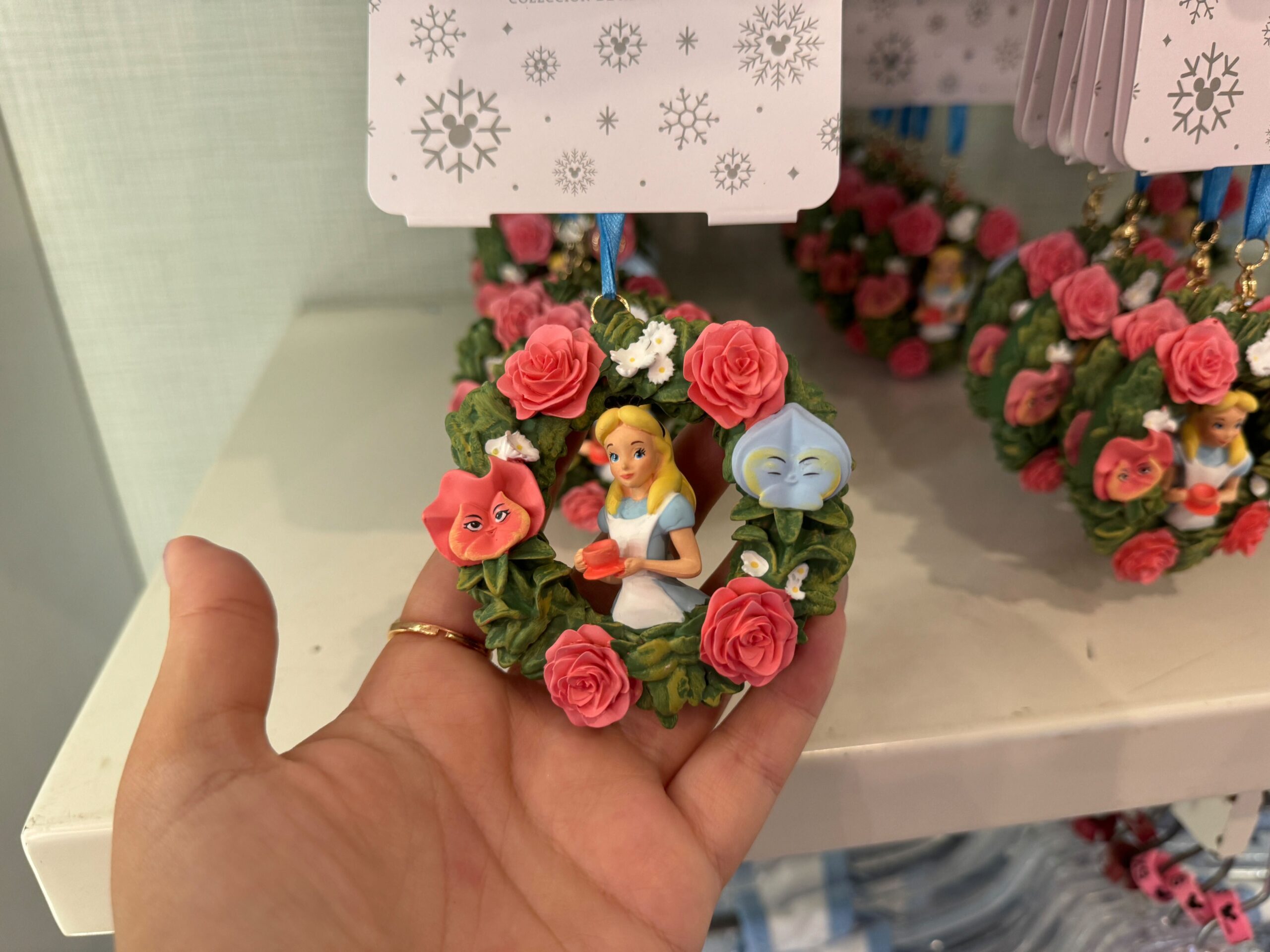 A hand holds a decorative ornament featuring a figure of a girl in a blue dress surrounded by flowers, displayed in a store.