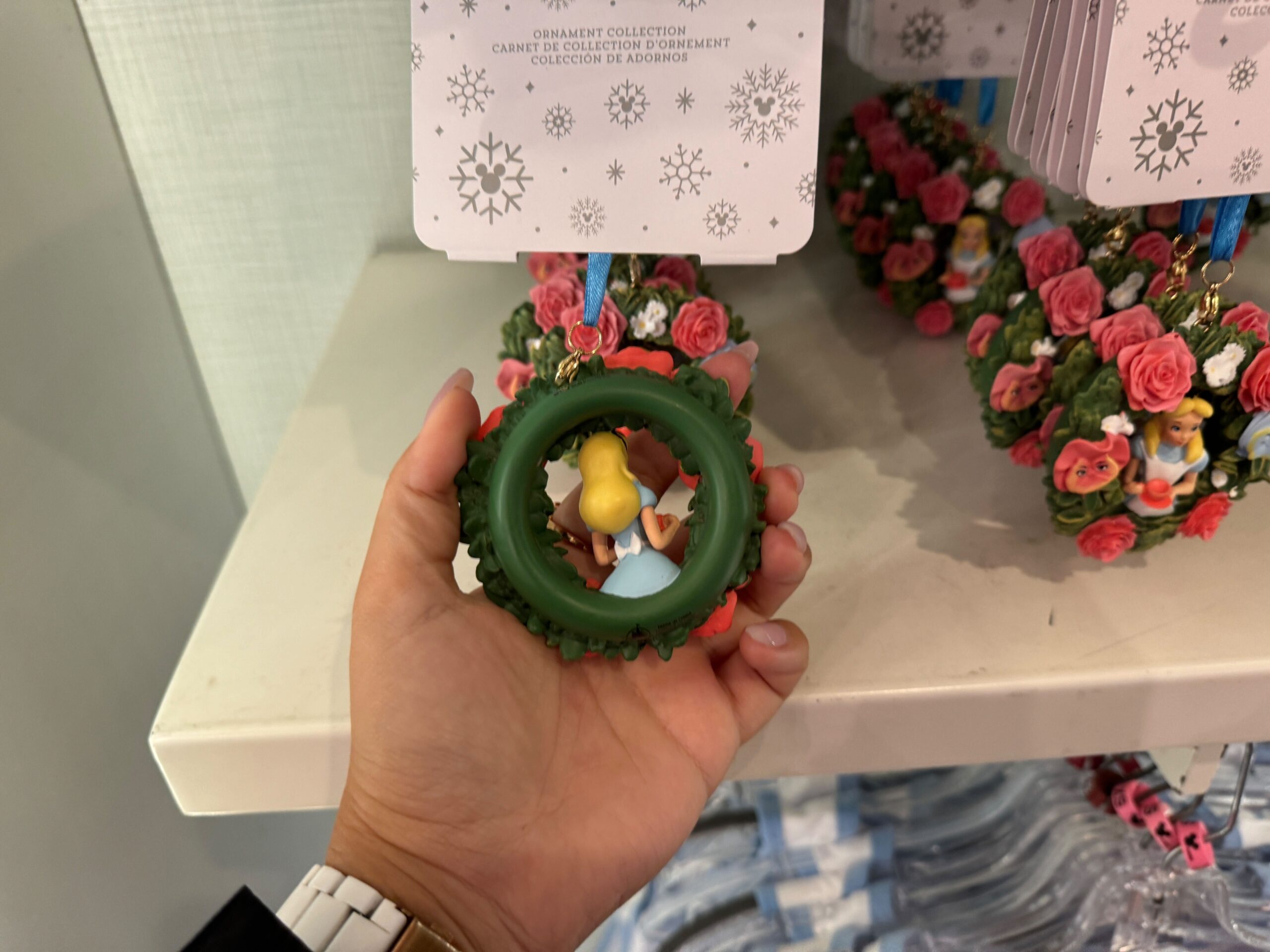 Hand holding a decorative wreath ornament featuring a yellow and blue figurine inside, displayed on a shelf with other floral ornaments.