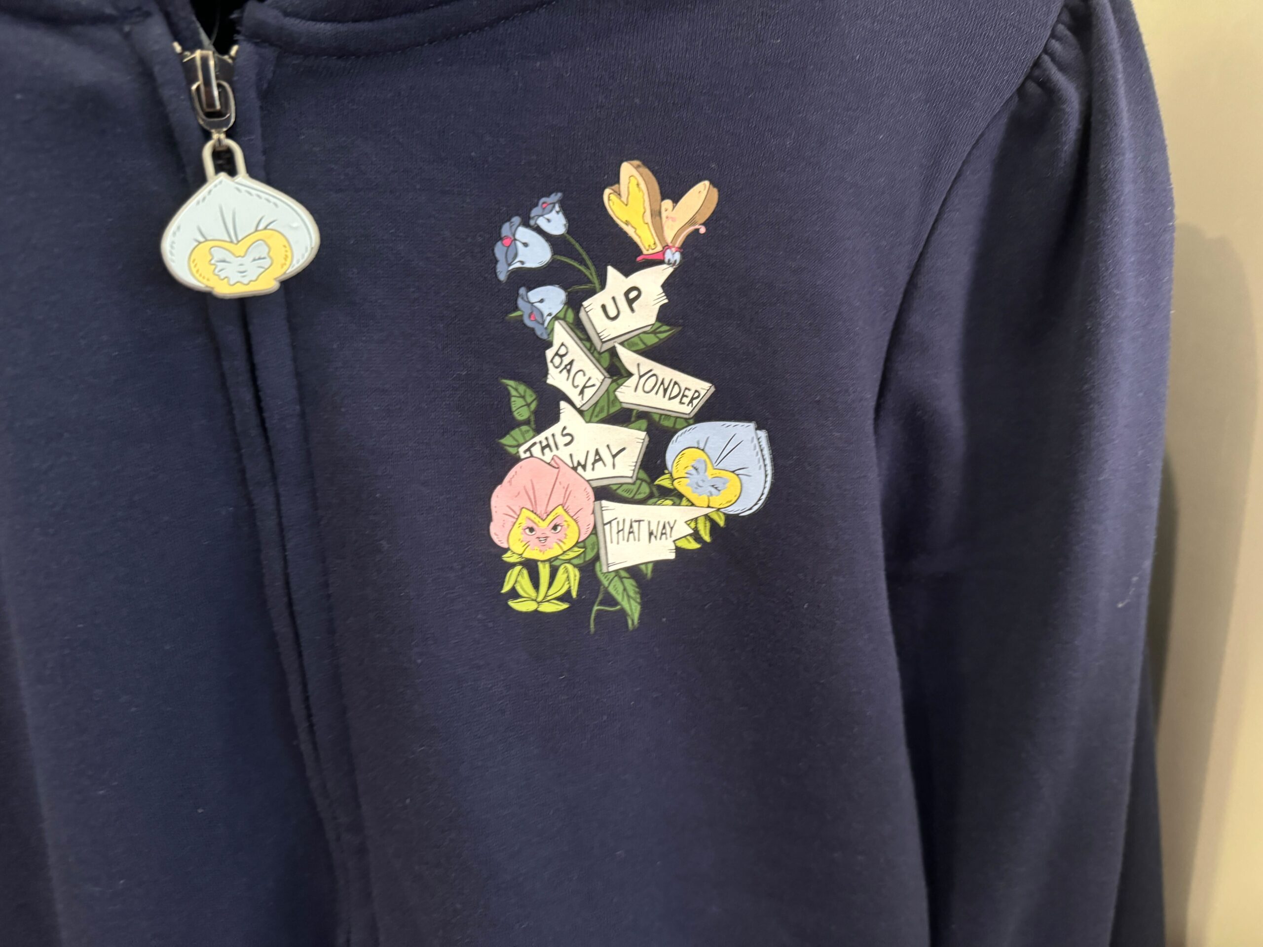 Close-up of a navy blue hoodie with an embroidered floral design and arrows. The text on the arrows reads "Up," "Down," "Back," "Yonder," "This Way," and "That Way.