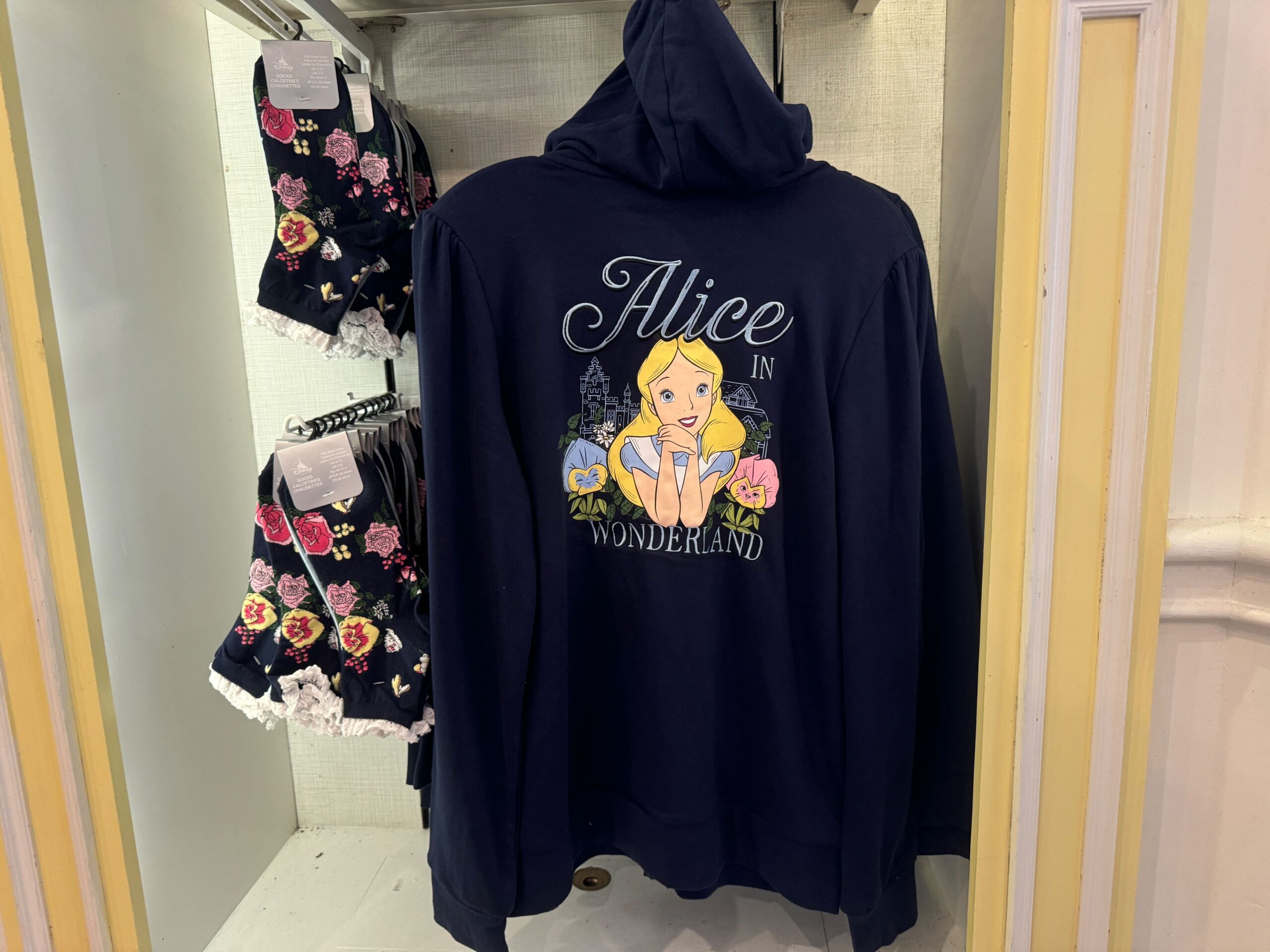 A navy hoodie with "Alice in Wonderland" text and character graphic on the back is displayed next to floral-patterned clothing on a rack.