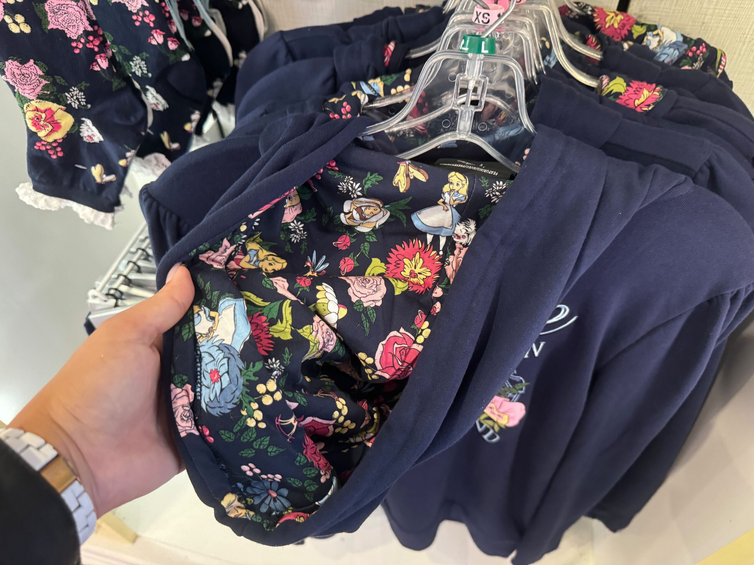 A person holding a blue jacket with a floral and character print lining, displayed on a clothing rack with similar jackets on hangers.