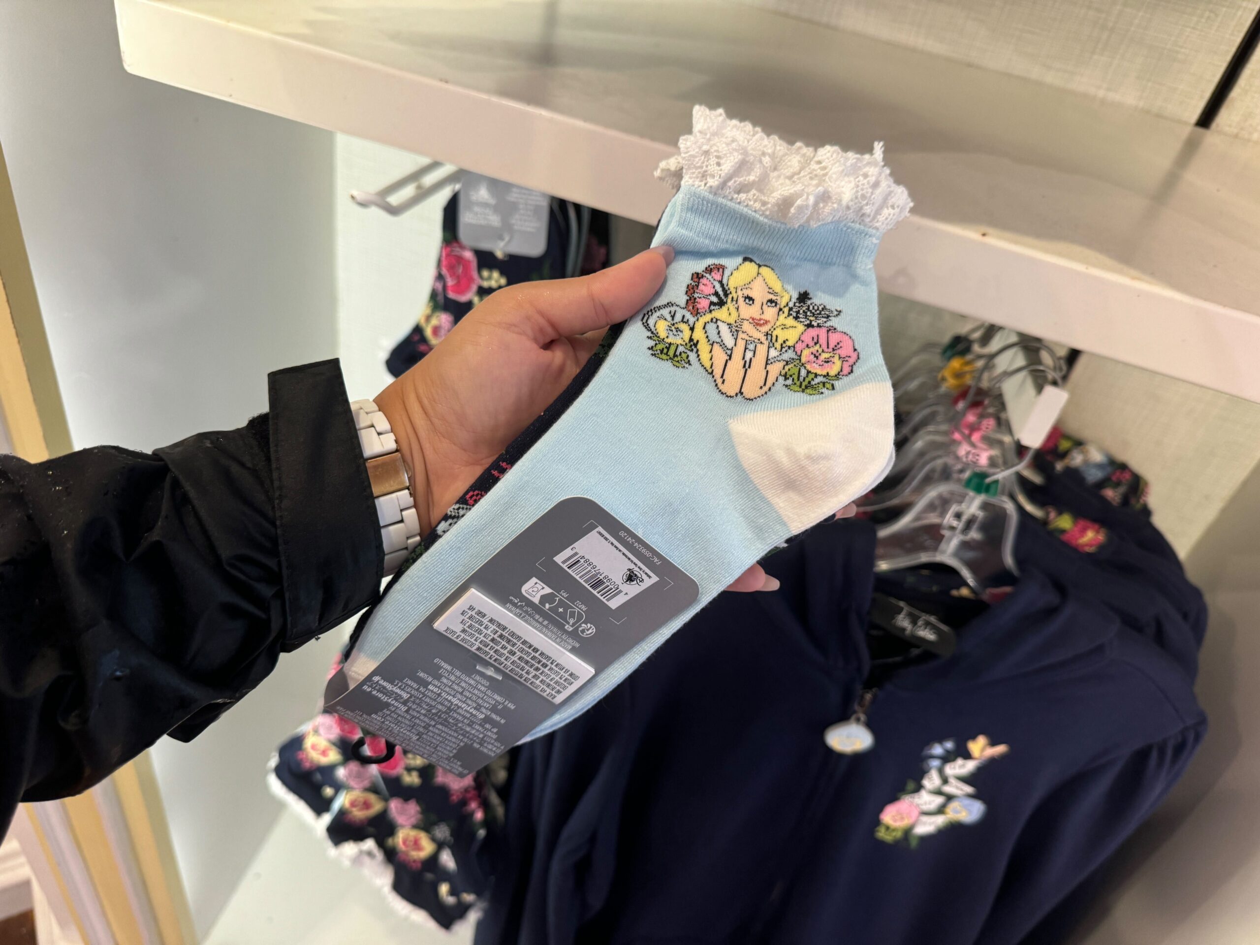 A hand holds a pair of socks with a floral and character design, near clothing with similar motifs.