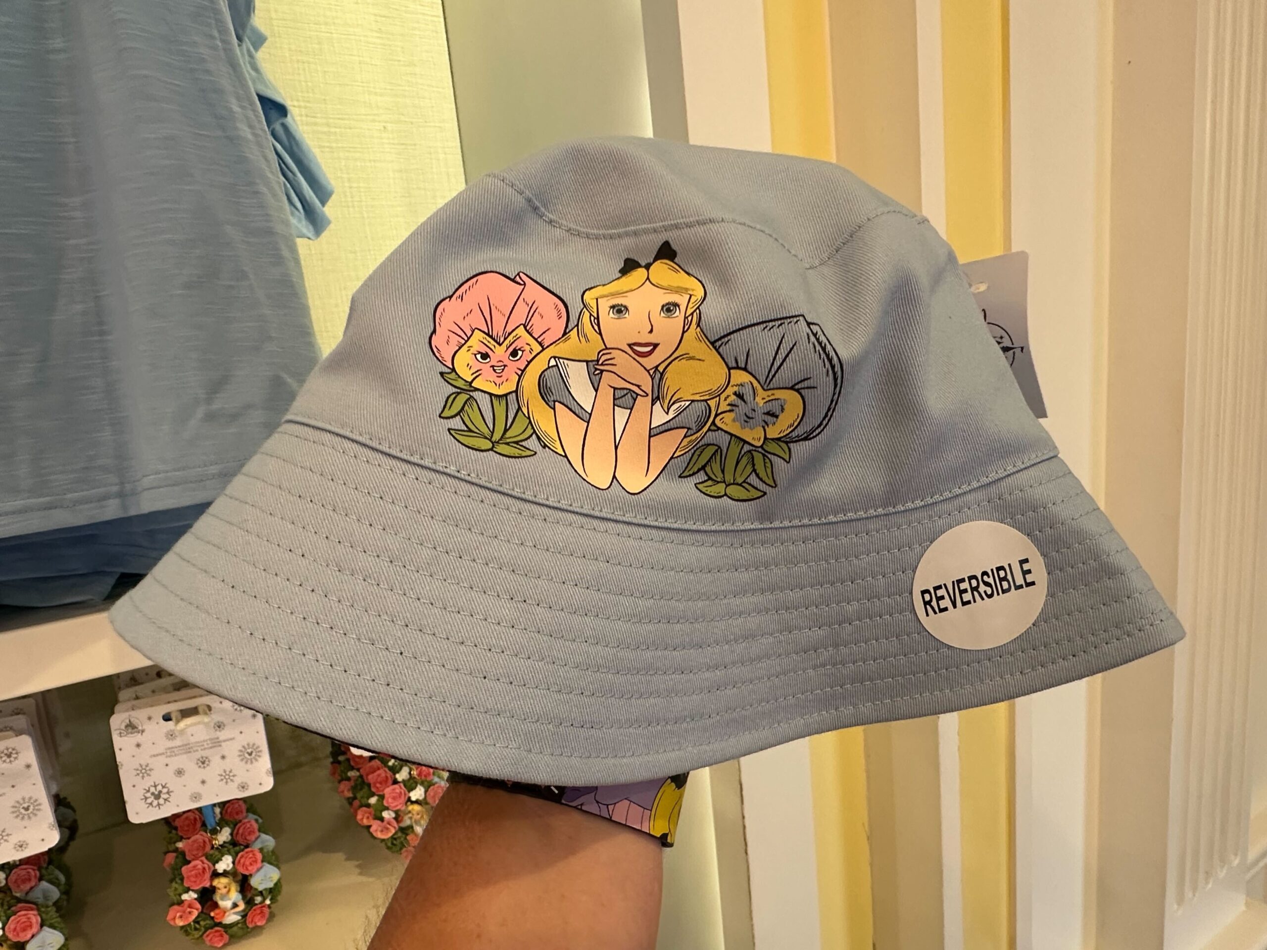 This light blue reversible bucket hat, part of the Alice in Wonderland merchandise collection, features a whimsical cartoon character surrounded by delightful flowers.