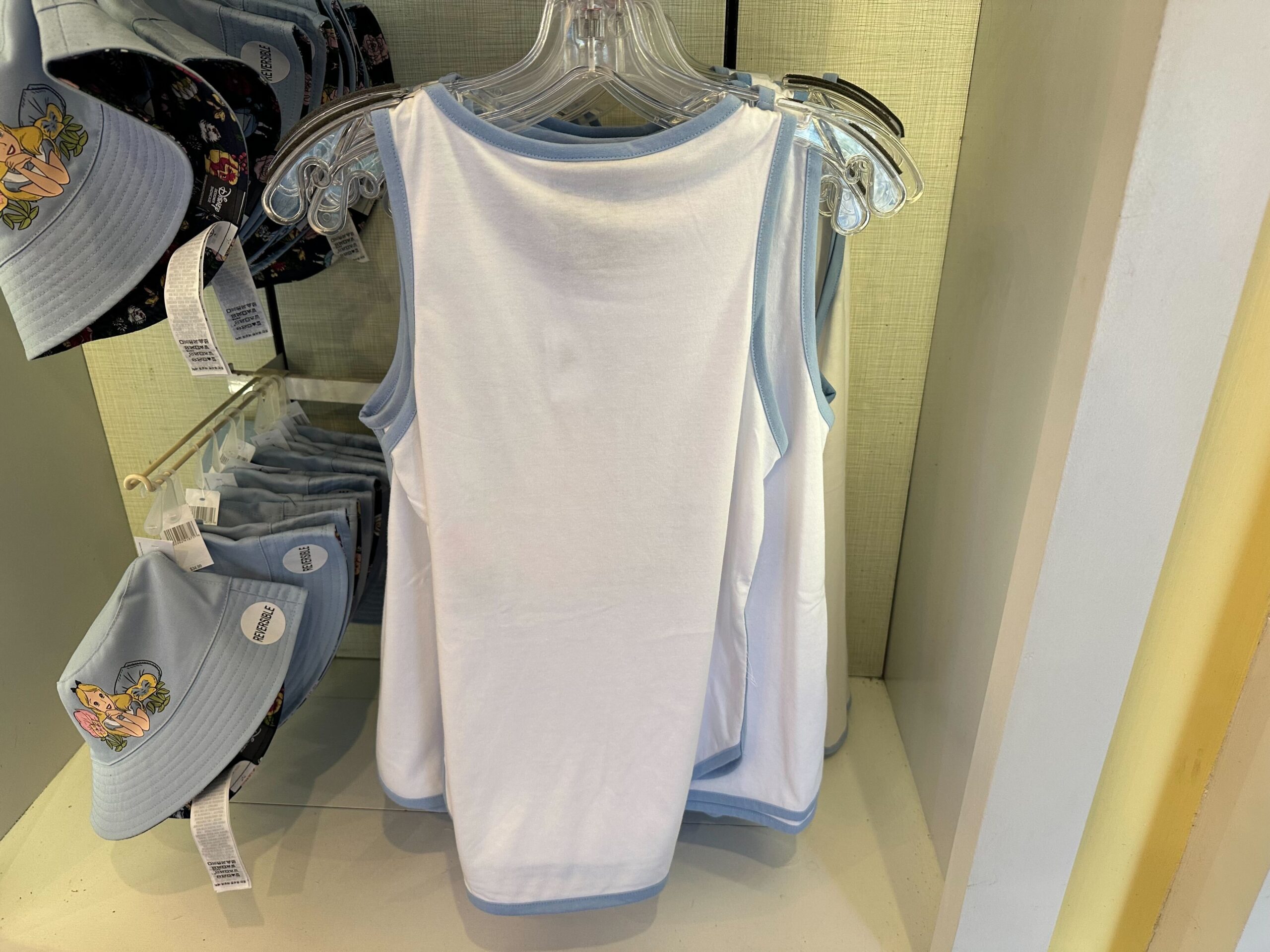 White and light blue tank tops on hangers in a store, placed next to a display of hats with whimsical cartoon designs, evoke a touch of Alice in Wonderland magic.