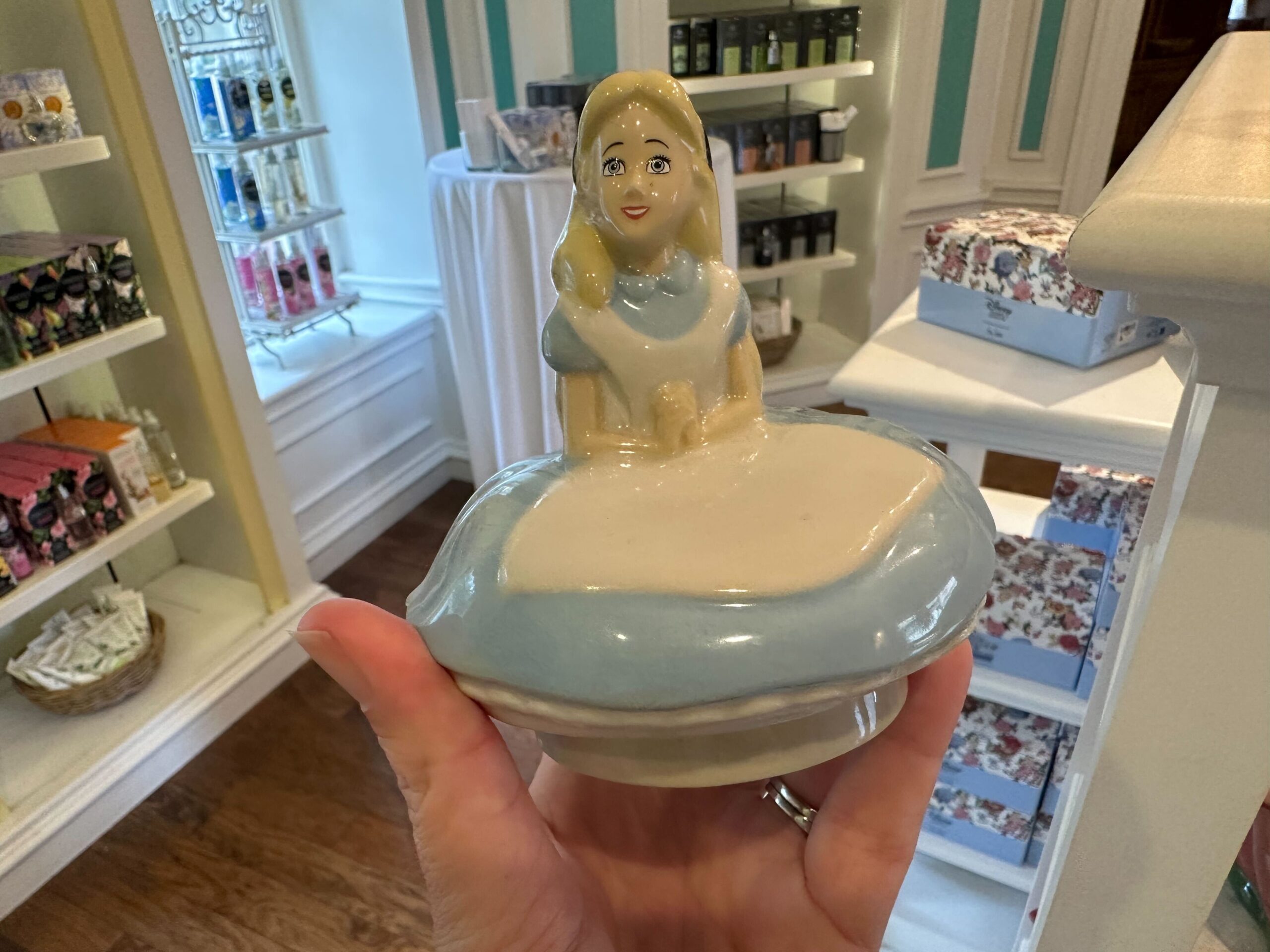 A ceramic container shaped like a figure in a blue and white dress, reminiscent of Alice in Wonderland, is held in a hand inside a store with shelves of merchandise in the background.