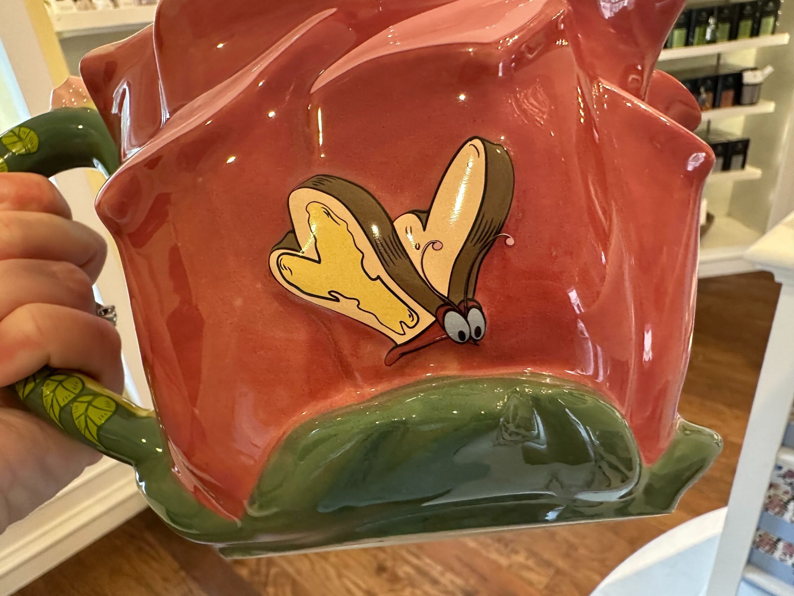 Close-up of a hand holding a red ceramic teapot featuring a whimsical cartoon butterfly design, reminiscent of Alice in Wonderland merchandise.