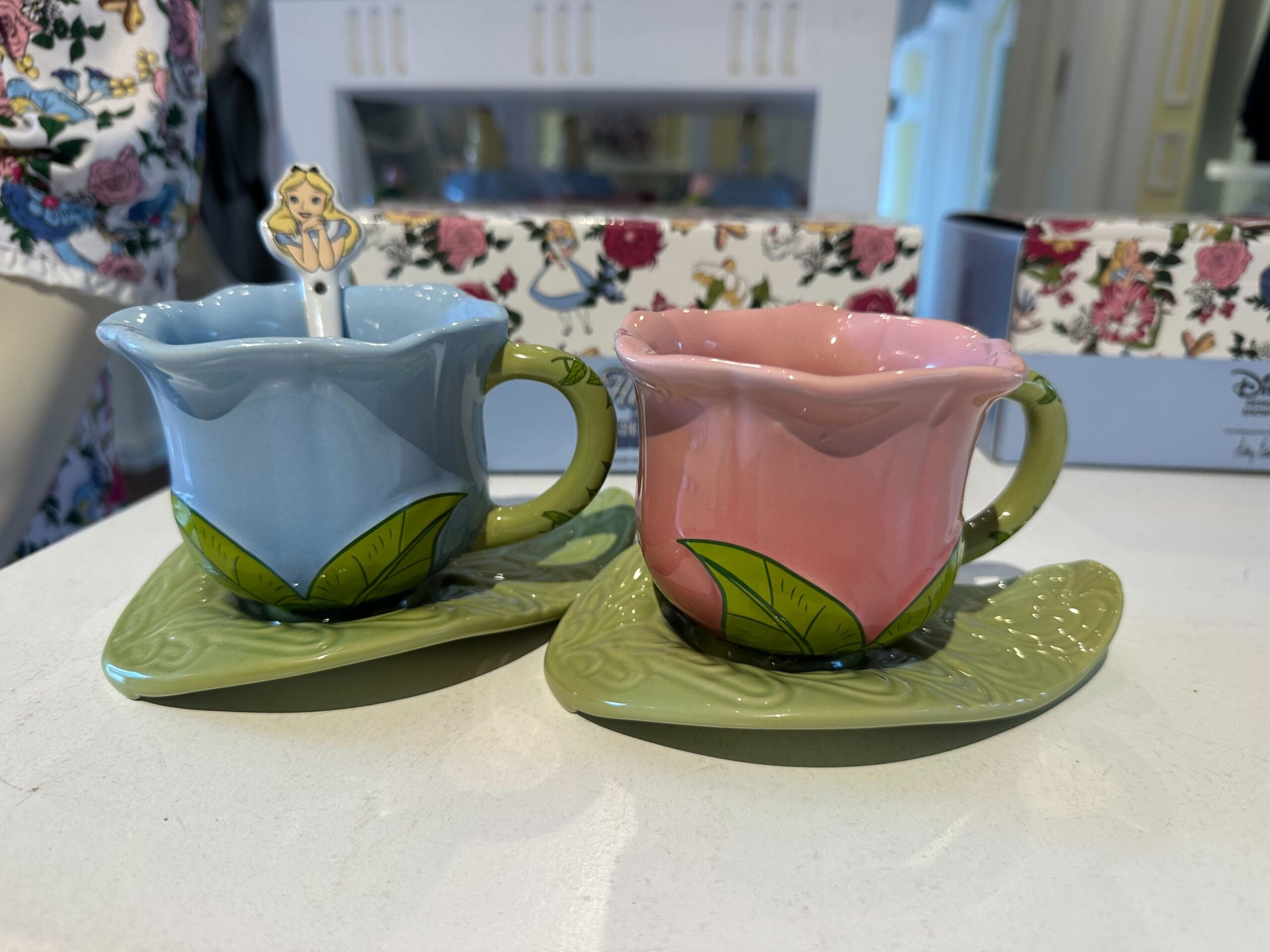 Two ceramic teacups shaped like flowers, one blue and one pink, each on a green leaf-shaped saucer. Reminiscent of Alice in Wonderland, a small stick figure decoration rests in the blue cup.