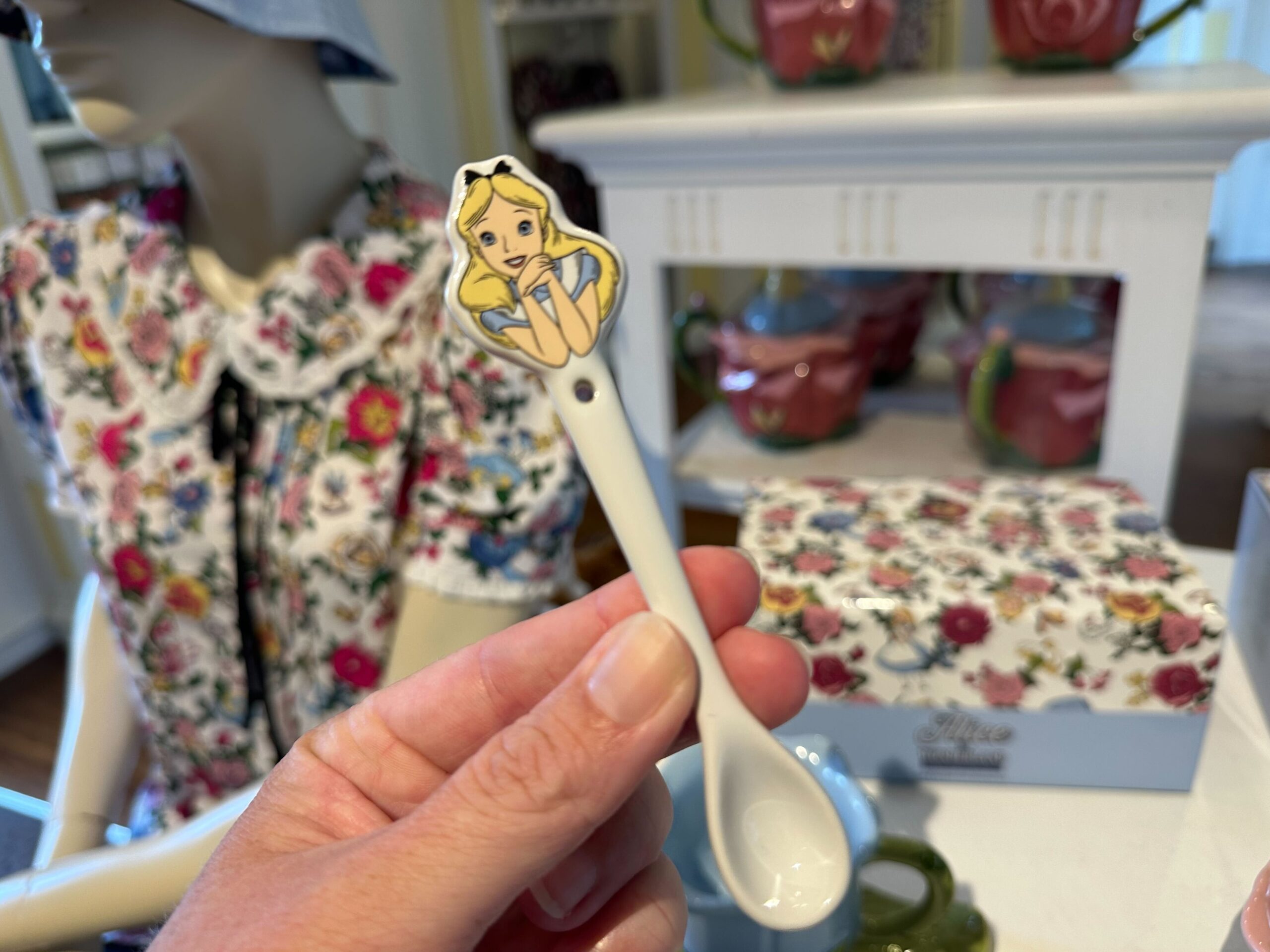 A hand holding a white spoon adorned with an animated girl reminiscent of Alice in Wonderland graces the scene, set against a backdrop of floral-patterned clothing and ceramic items.