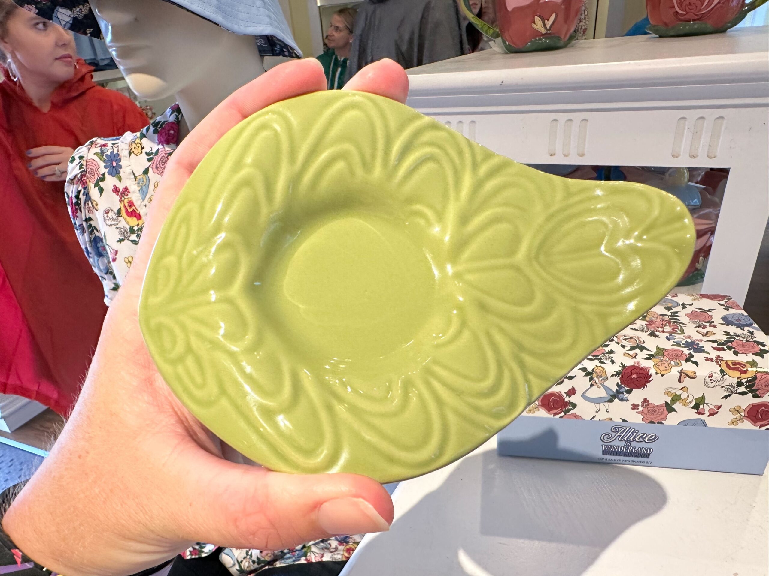 A hand holding a green ceramic dish with an embossed leaf pattern, reminiscent of Alice in Wonderland, is displayed indoors.