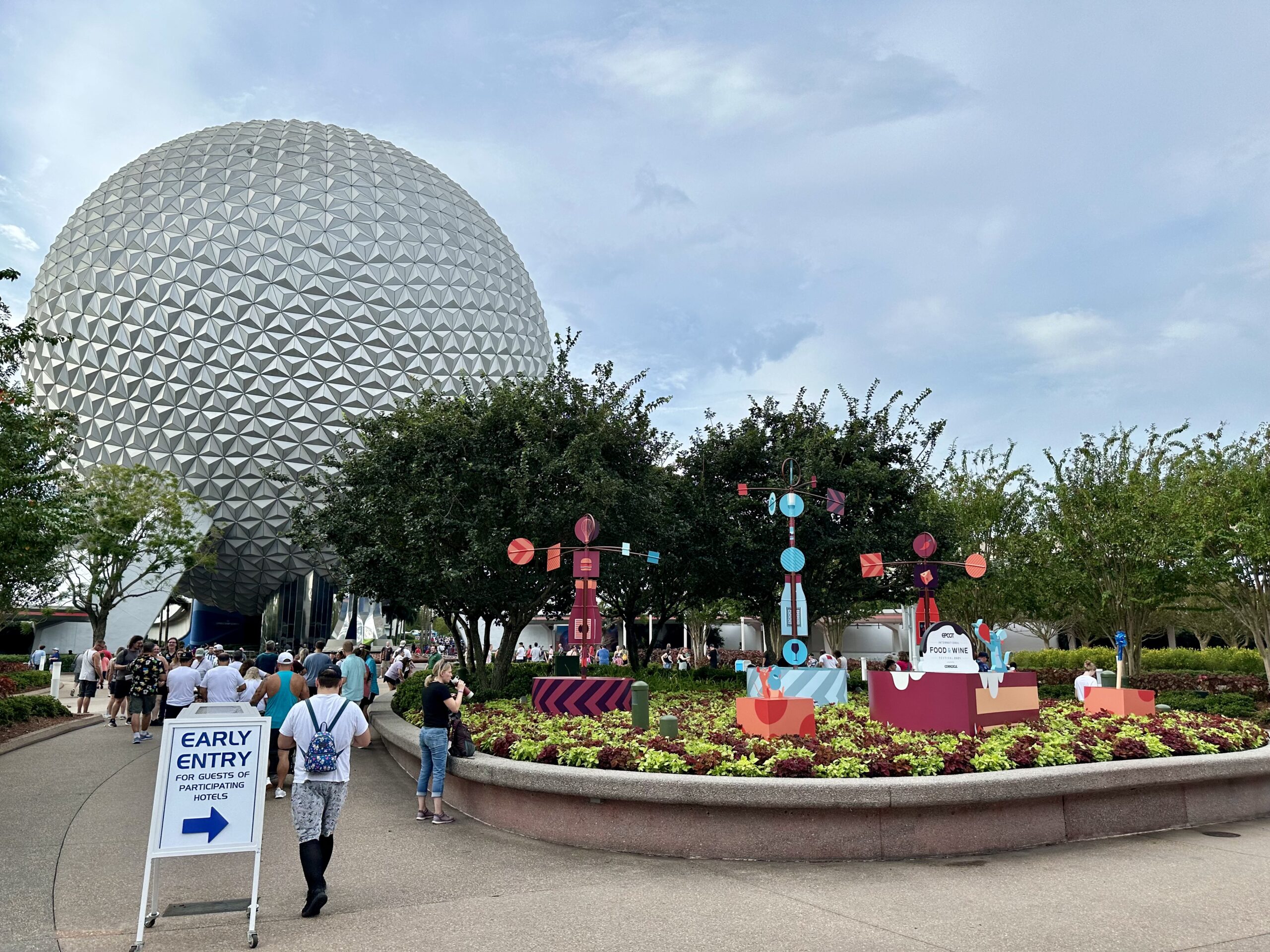 People walking towards a large geodesic dome structure with signs and decorations nearby. An "Early Entry for Guests" sign with an arrow is in the foreground. Trees and cloudy sky in the background.