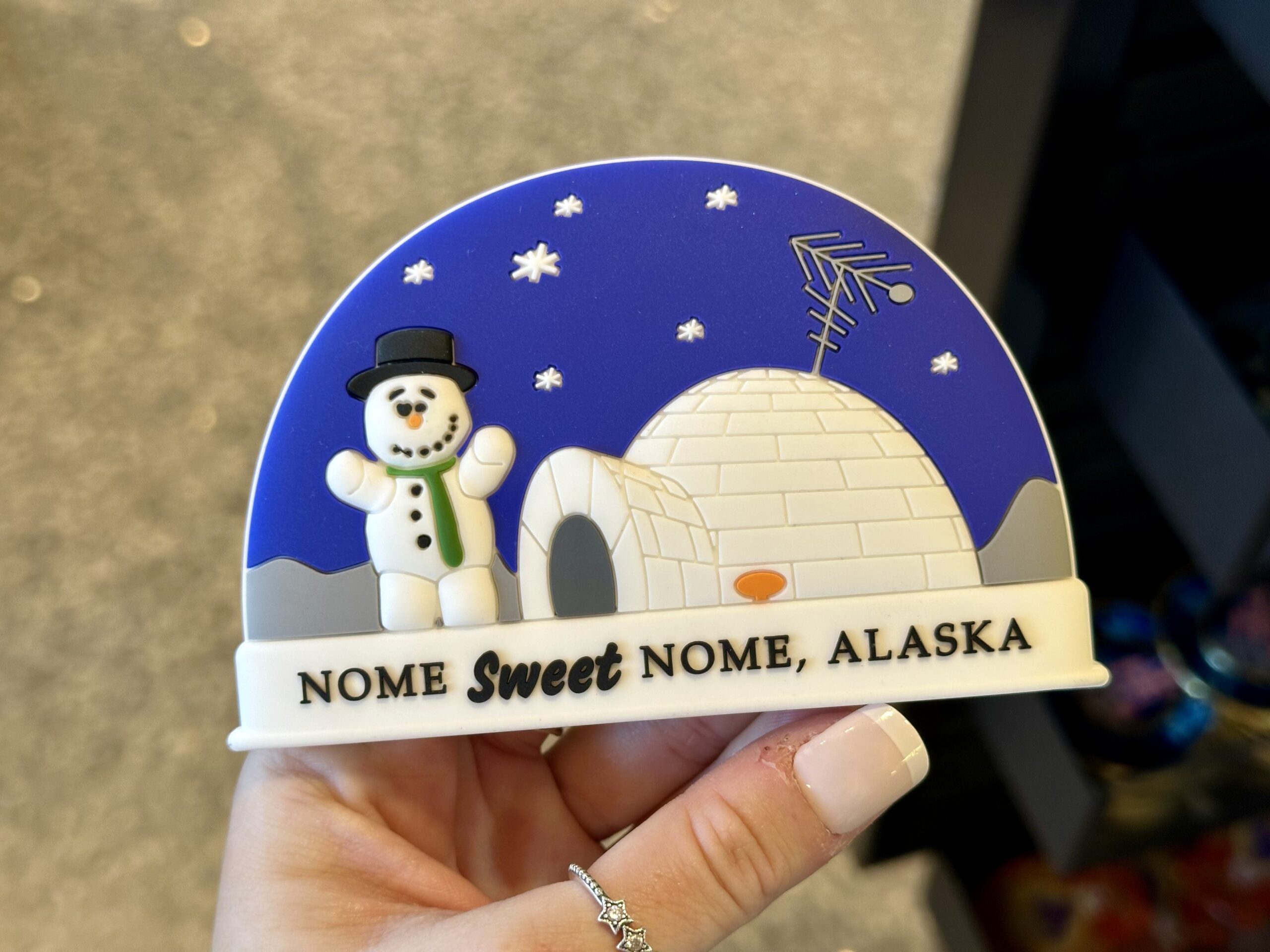 A person holds a magnet featuring a snowman, igloo, and satellite dish, with the text "NOME Sweet NOME, ALASKA" against a starry night background.