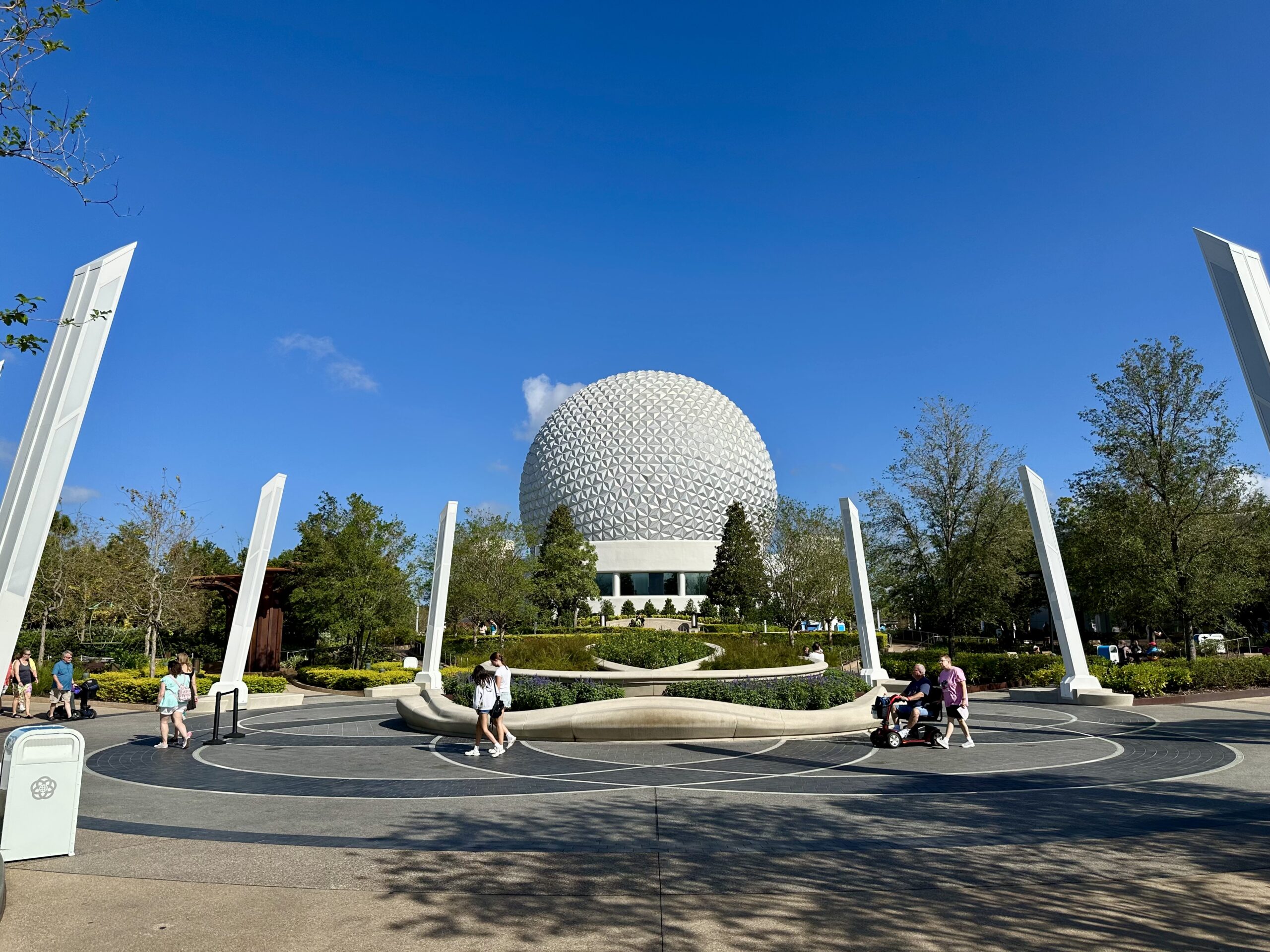 A large geodesic sphere at a theme park, surrounded by people walking and trees under a clear blue sky.
