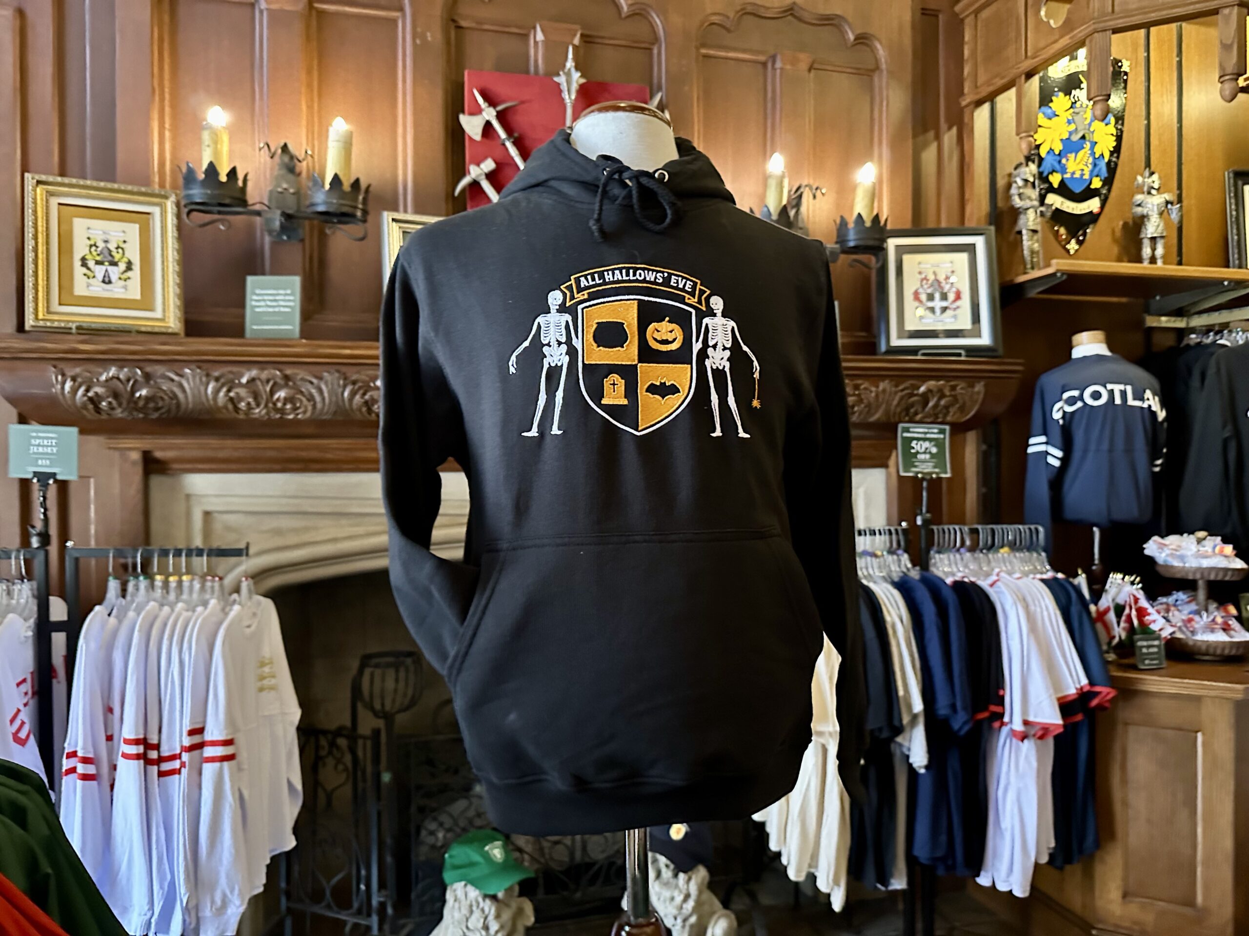 A black hoodie with a Halloween-themed crest design is displayed on a mannequin in a clothing store. Shirts hang in the background.