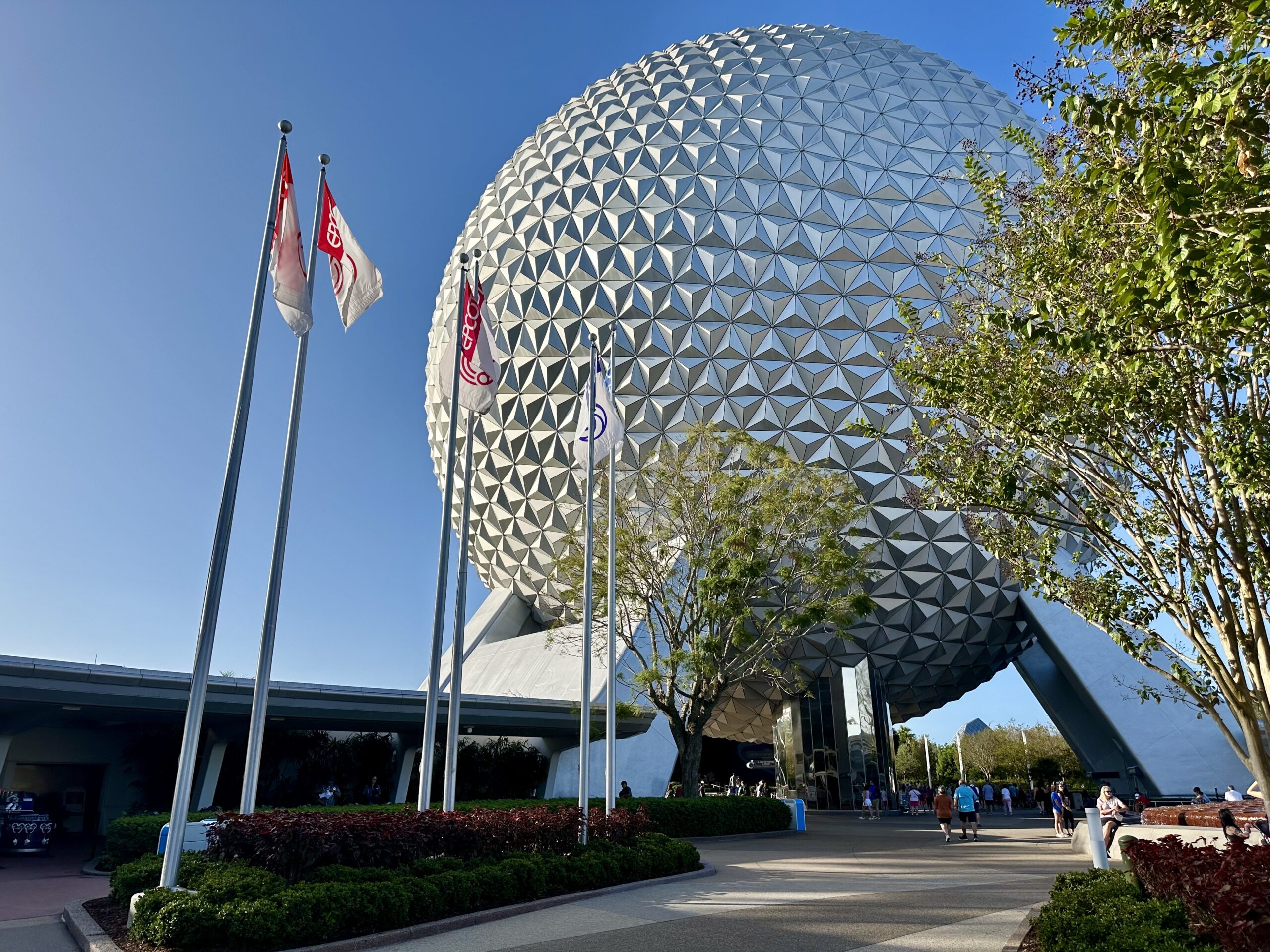 A large geodesic sphere with a pattern of triangular panels at an entrance, surrounded by flags and trees under a clear blue sky.