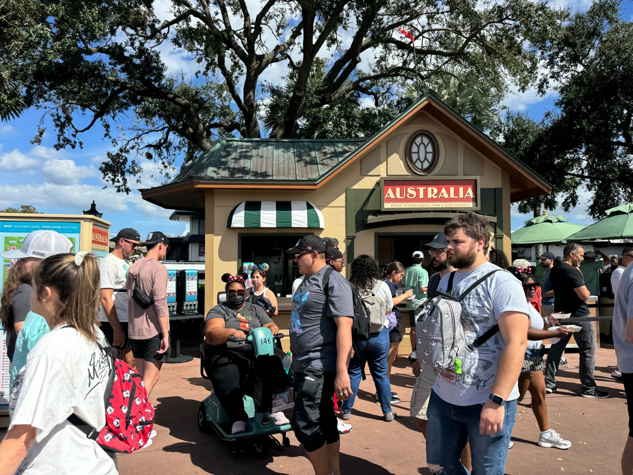 A crowd gathered outside the "Australia" building, with trees and a clear sky in the background, as part of EPCOT's vibrant Food & Wine Festival. People are walking and standing around, exploring each unique booth.