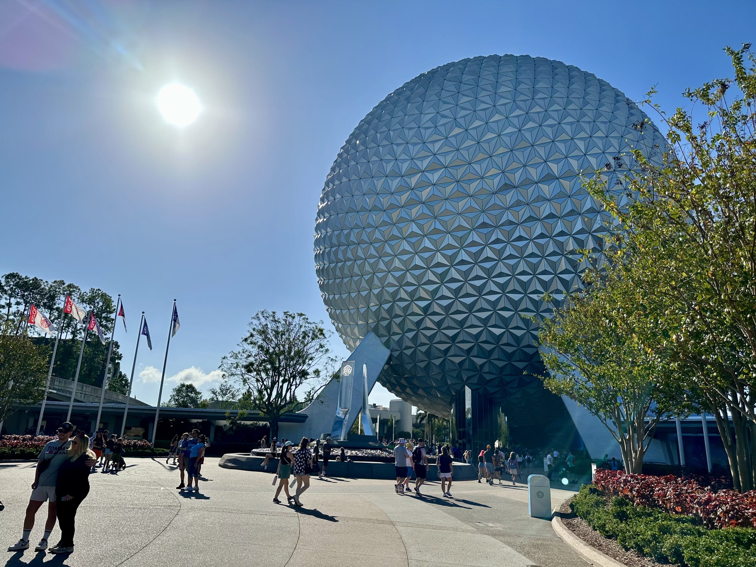 Visitors walk near a large geodesic sphere under a clear blue sky with the sun shining brightly. Flags and trees are visible in the background.