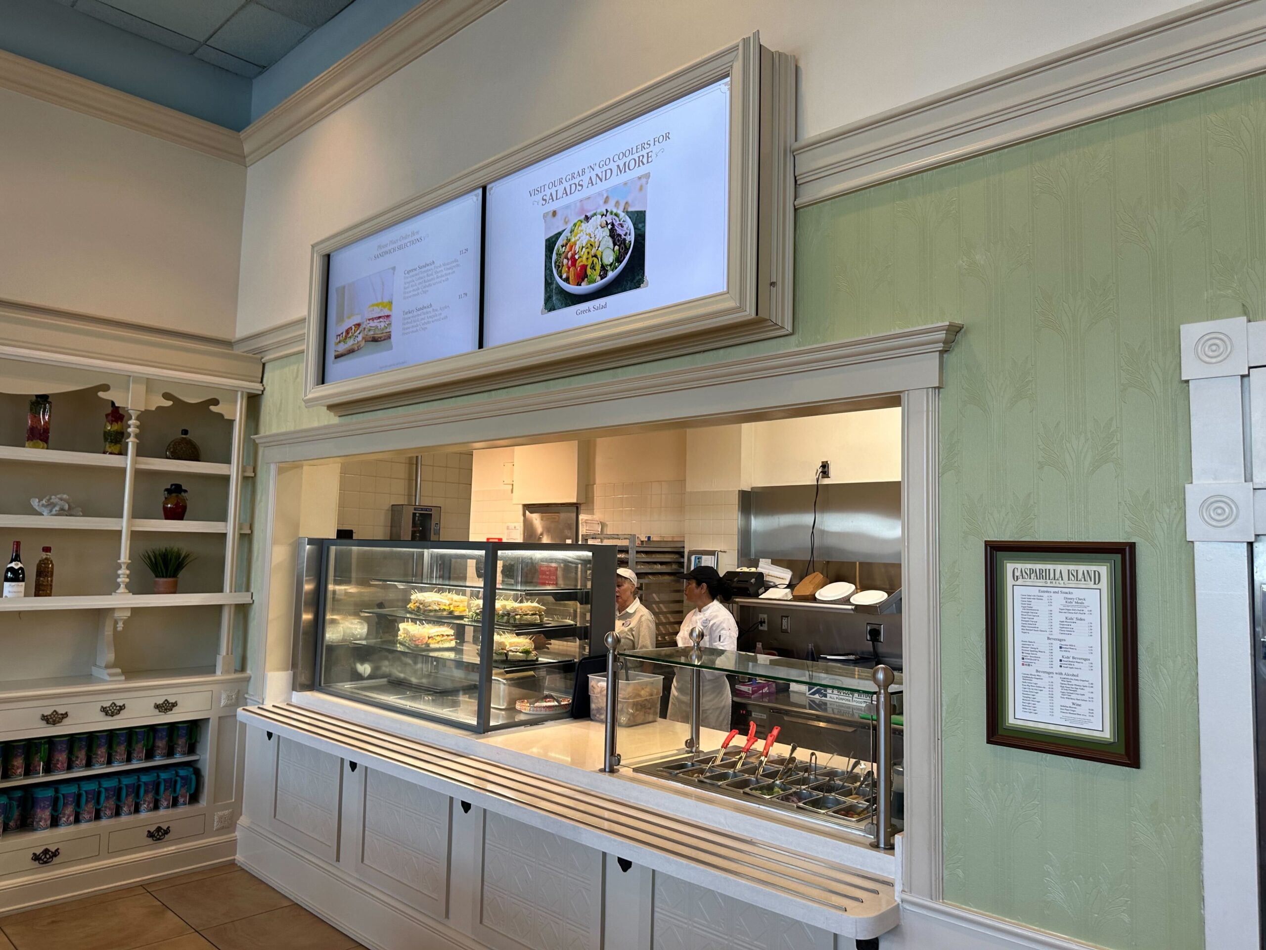 A restaurant counter with two employees working in the kitchen. Above the counter are two display screens showing menu items. Shelves with condiments and a menu board are on the right wall.