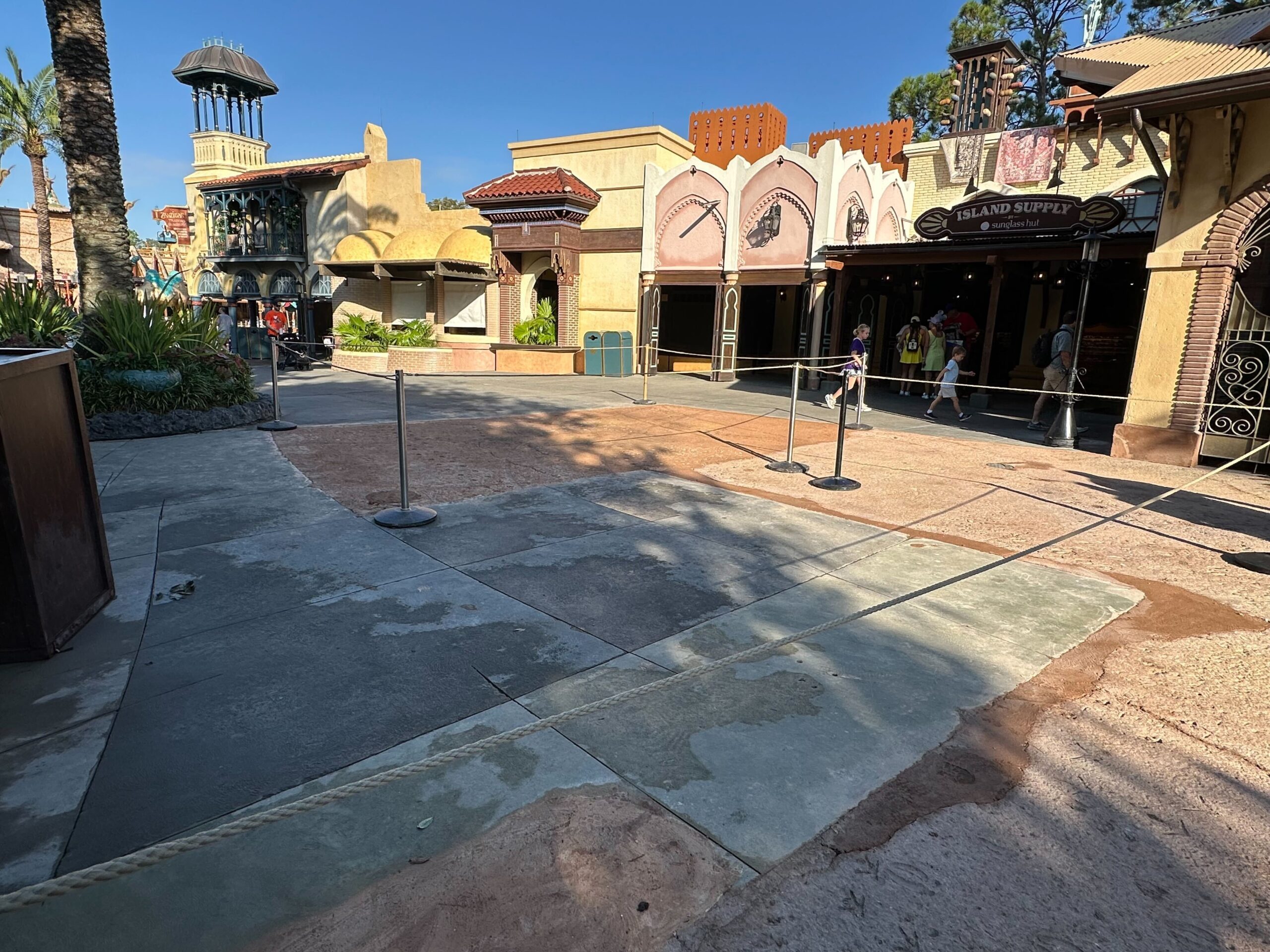 An empty outdoor plaza with themed buildings, palm trees, and some people in the background. Ropes and metal stanchions are set up on the pavement.