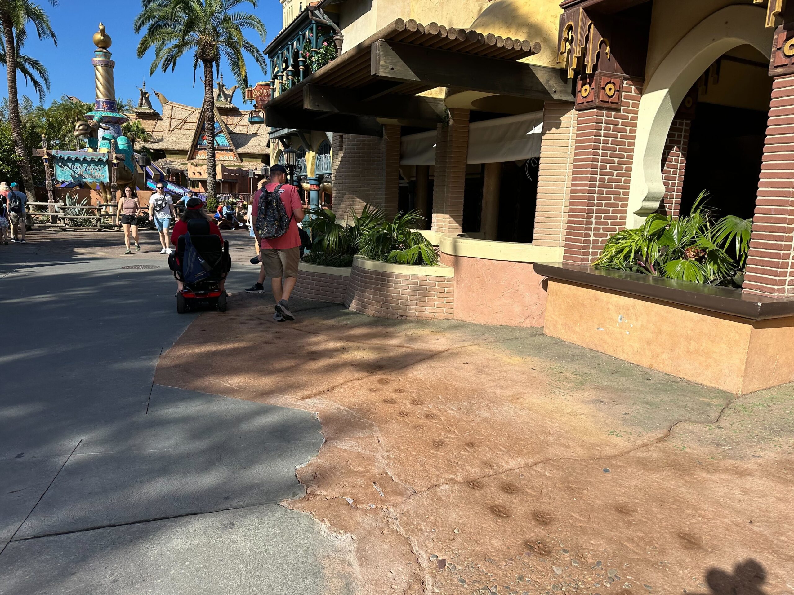 A person in a red shirt walks and another uses a mobility scooter on a sunny path bordered by themed buildings with palm trees.