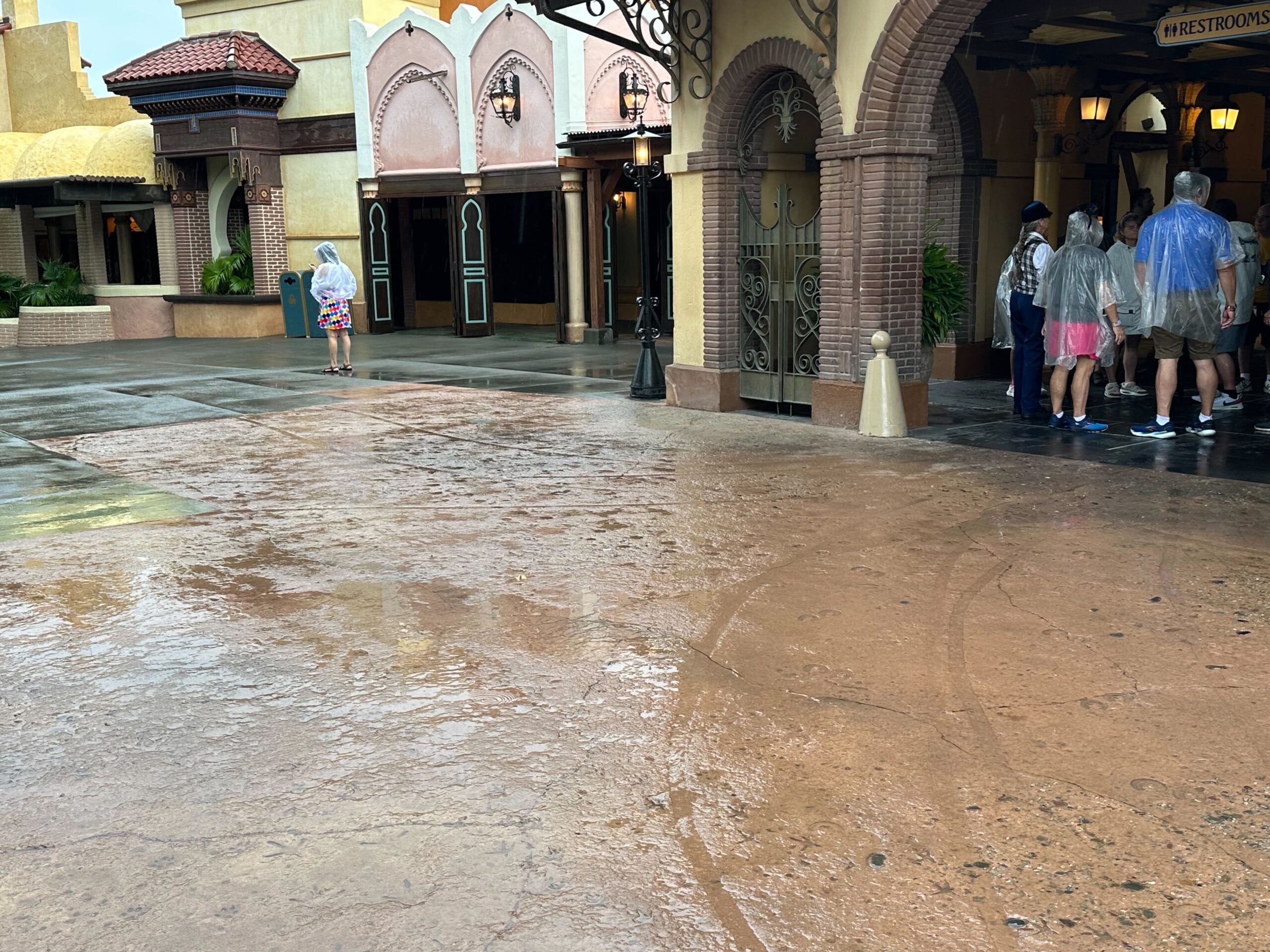 People wearing rain ponchos stand near the restrooms in a theme park area on a rainy day. Wet pavement shows visible tire tracks.