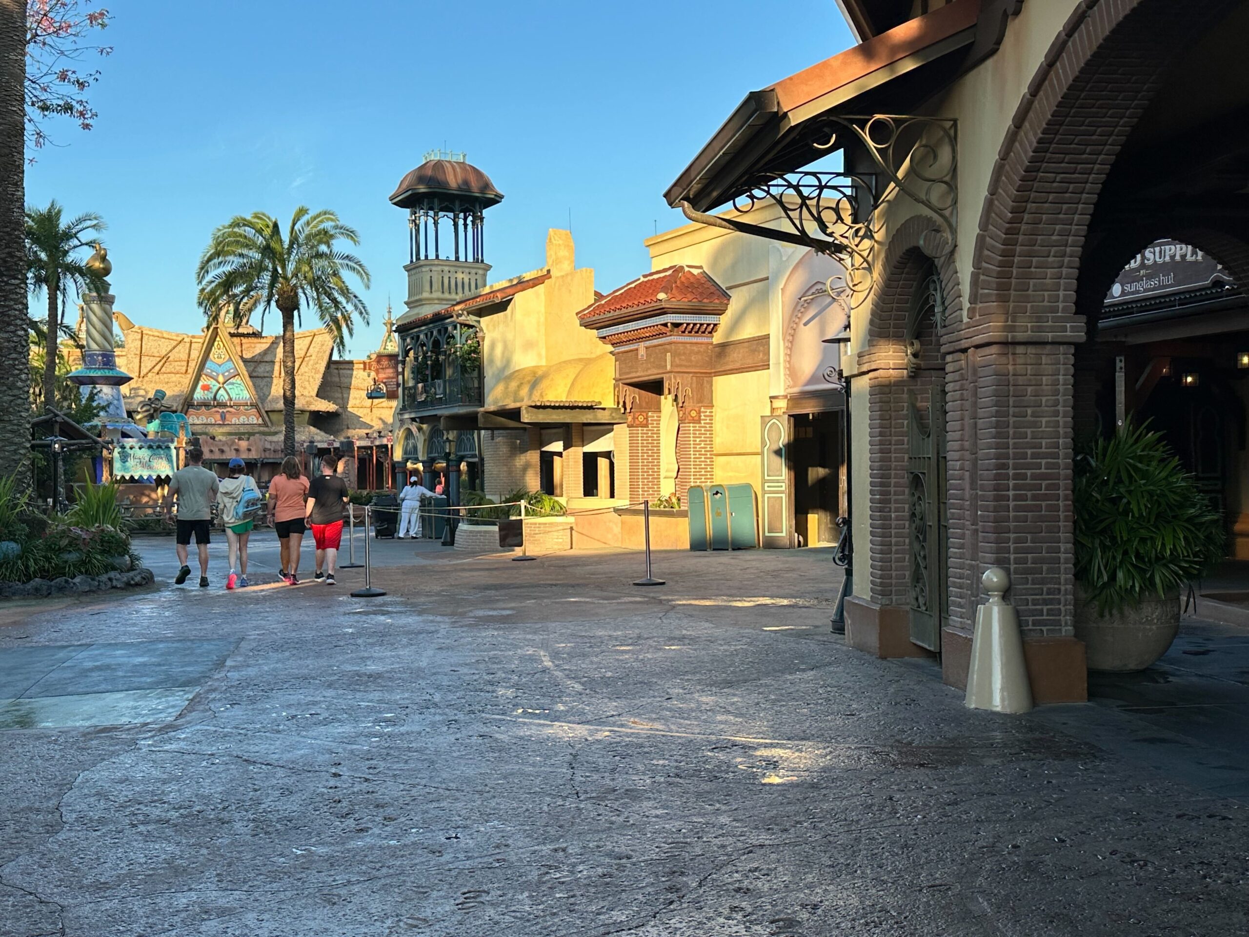 A group of people walk through a theme park with colorful buildings, palm trees, and clear blue sky.