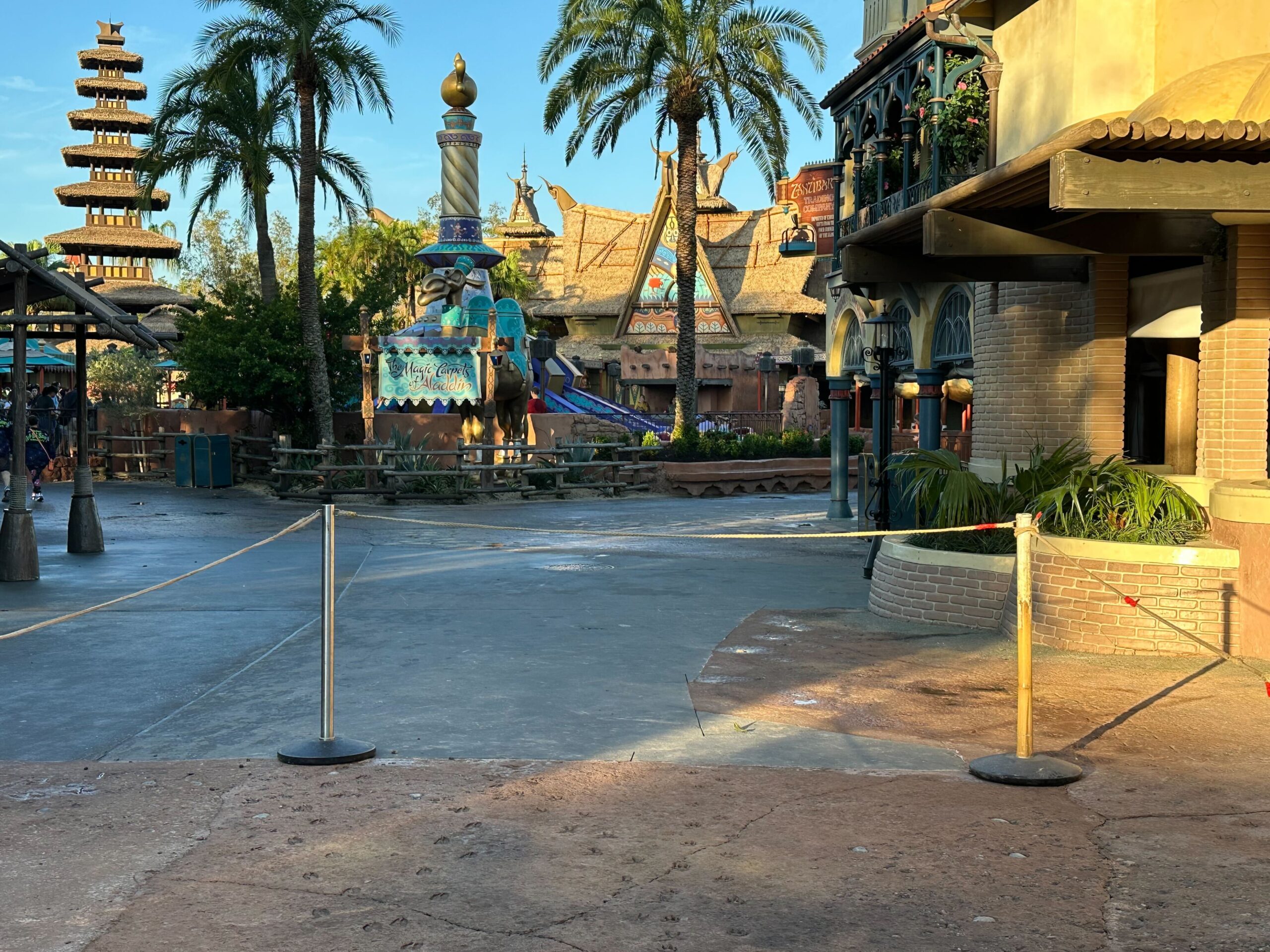 The image shows a rope barrier blocking access to a closed area in an amusement park with palm trees and themed architecture in the background.