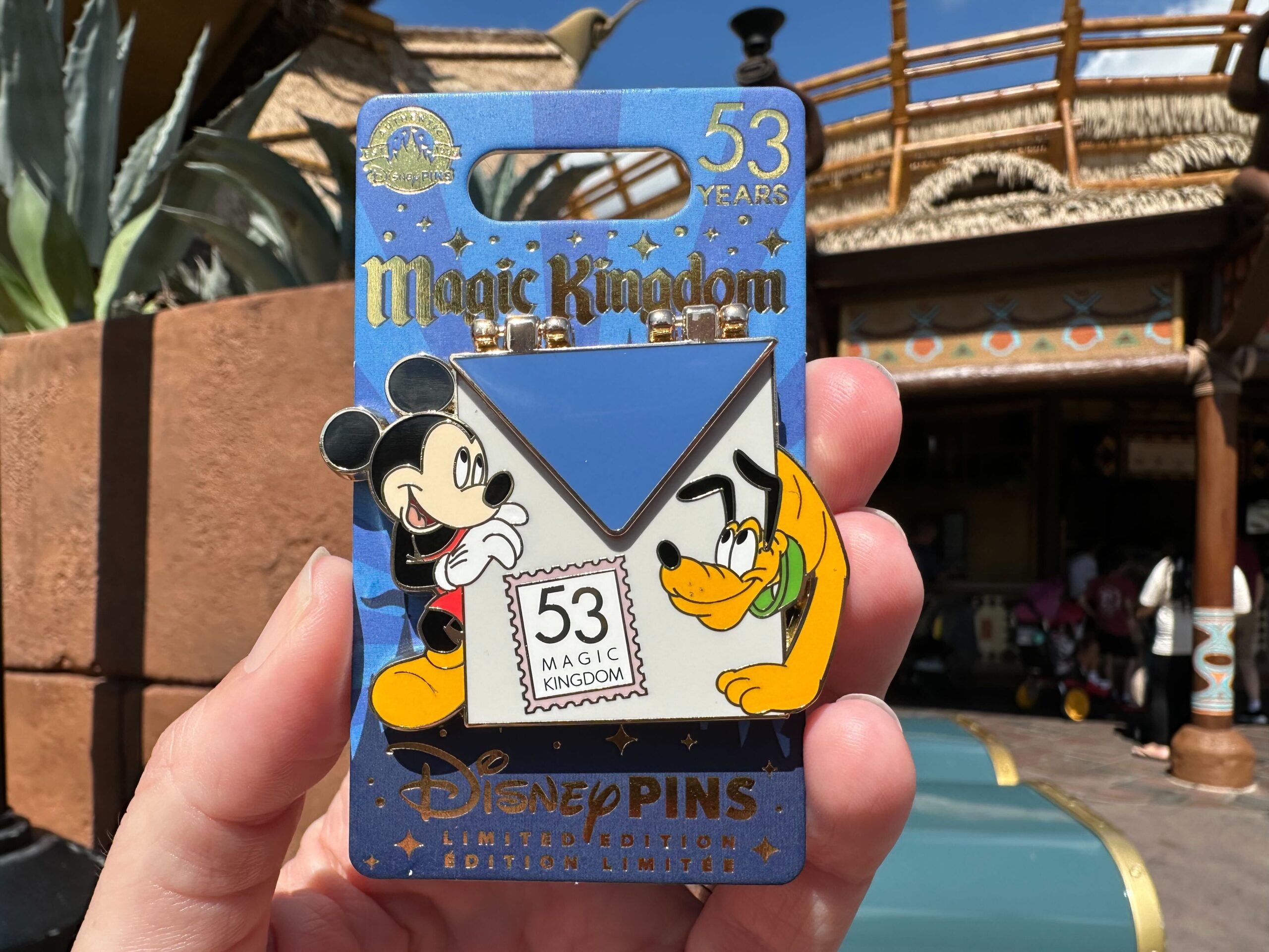 Hand holding a limited edition Disney pin featuring Mickey Mouse and Pluto celebrating 53 years of Magic Kingdom, with a background of a theme park attraction.
